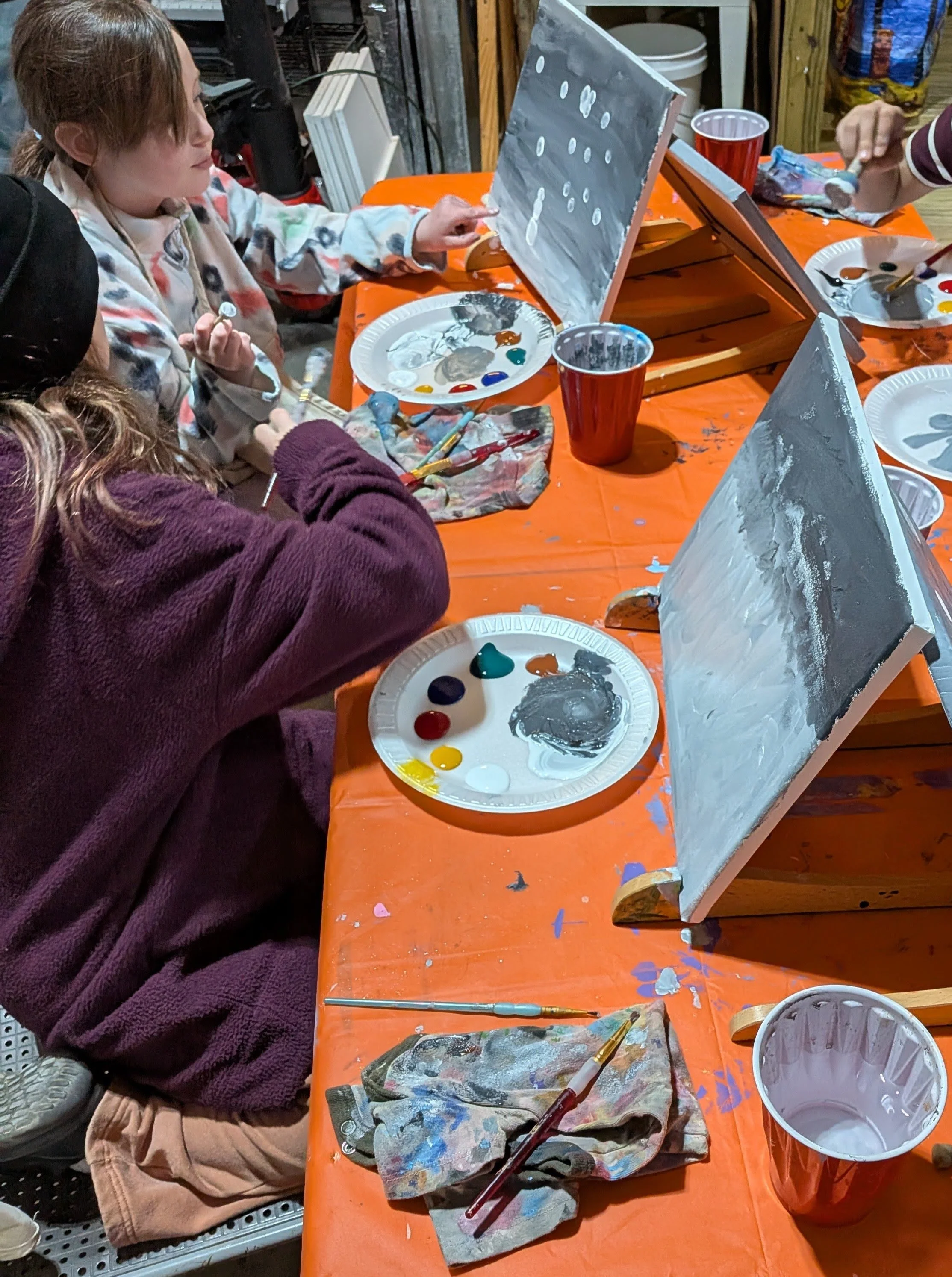 Children painting on canvases with gray and white color, seated at a table covered with an orange tablecloth, with paint palettes, brushes, and cups.