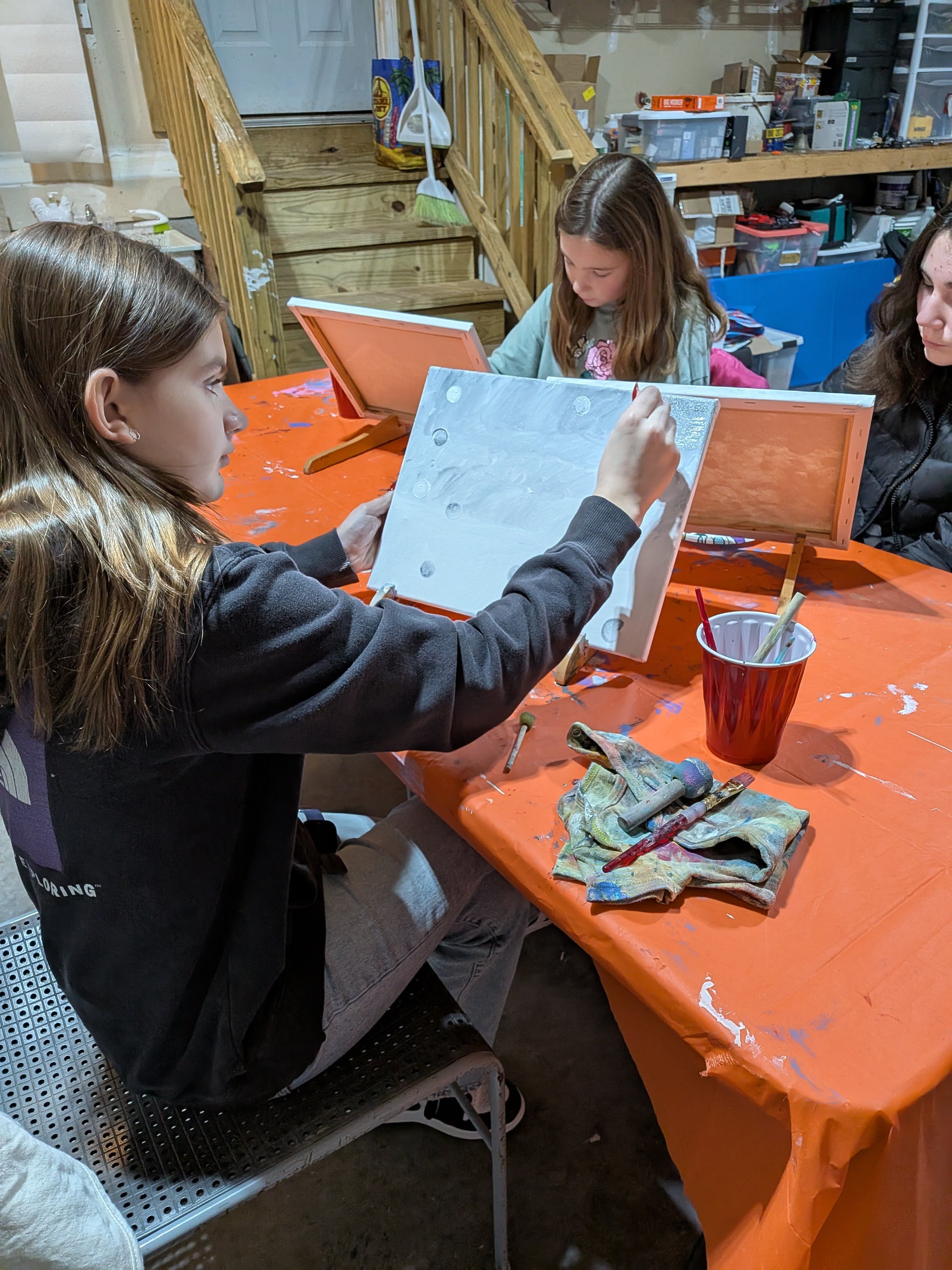 Group of children painting pottery at a table in a craft studio, with painting supplies and stored boxes in the background.