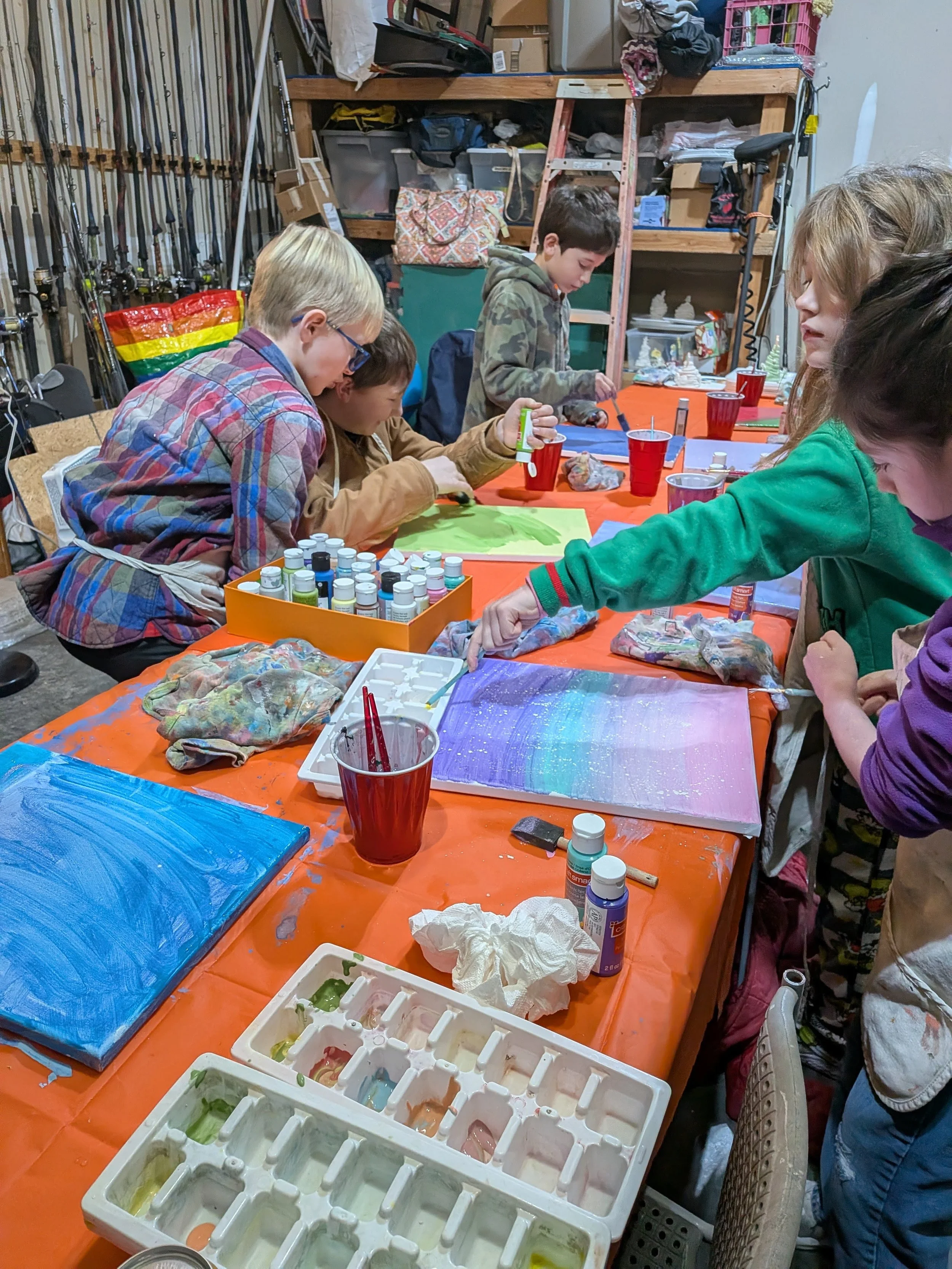 Children painting on canvases at a table covered with orange tablecloths, in a garage or craft room setting with various art supplies and paint bottles.