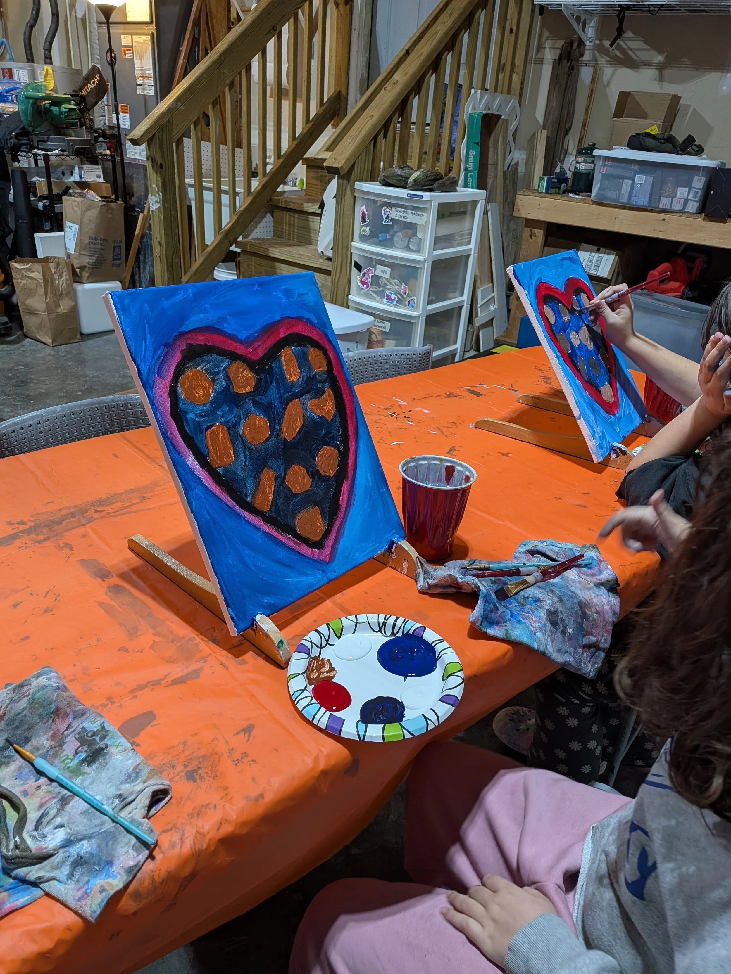 Children painting colorful heart-shaped artwork on canvases in a workshop with orange tablecloth, paintbrushes, palettes, and art supplies.