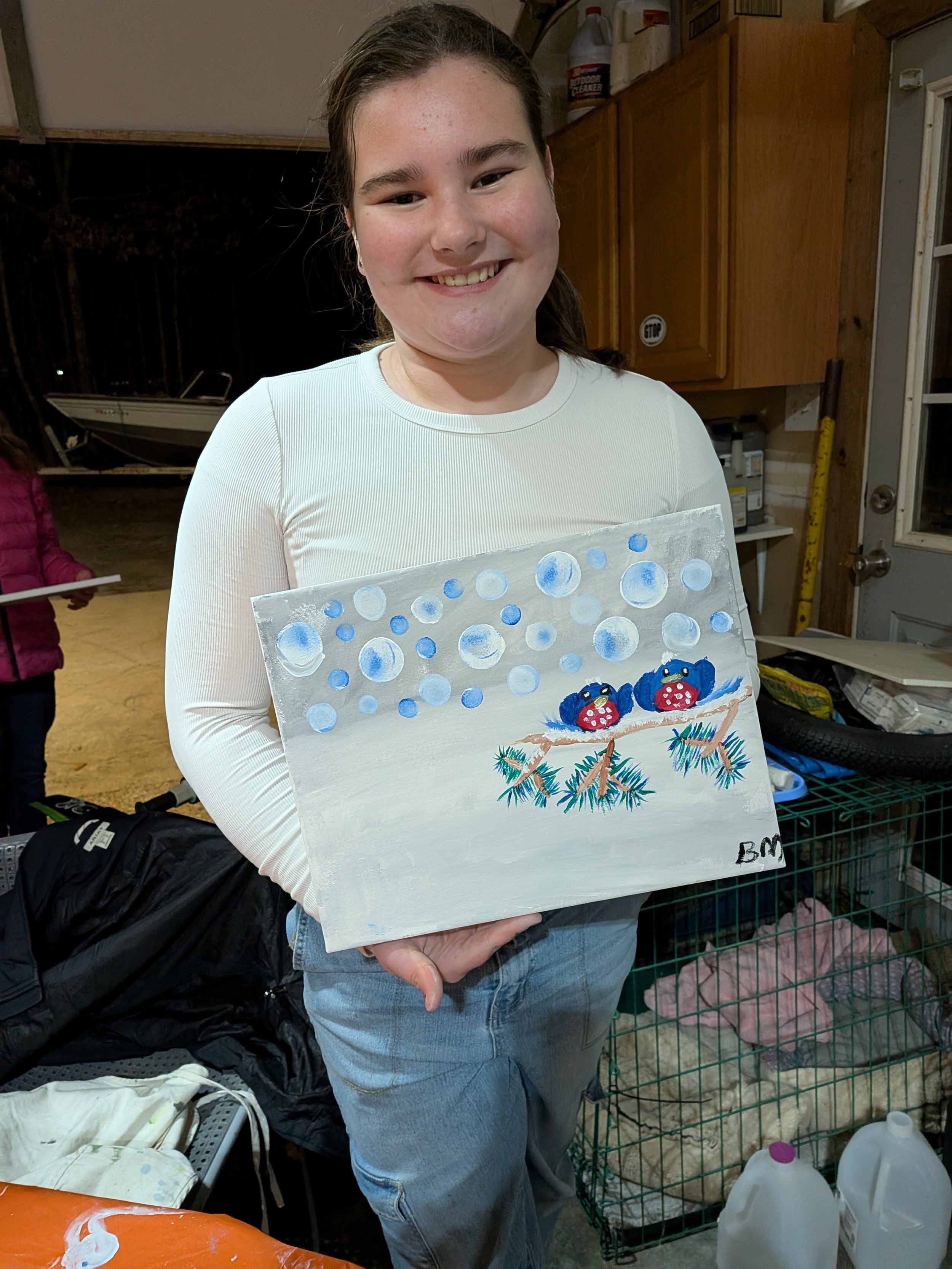 A young girl smiling and holding a painting with blue, white, and red ladybugs on a branch, and a snowy background with snowballs, in a garage or workshop setting.