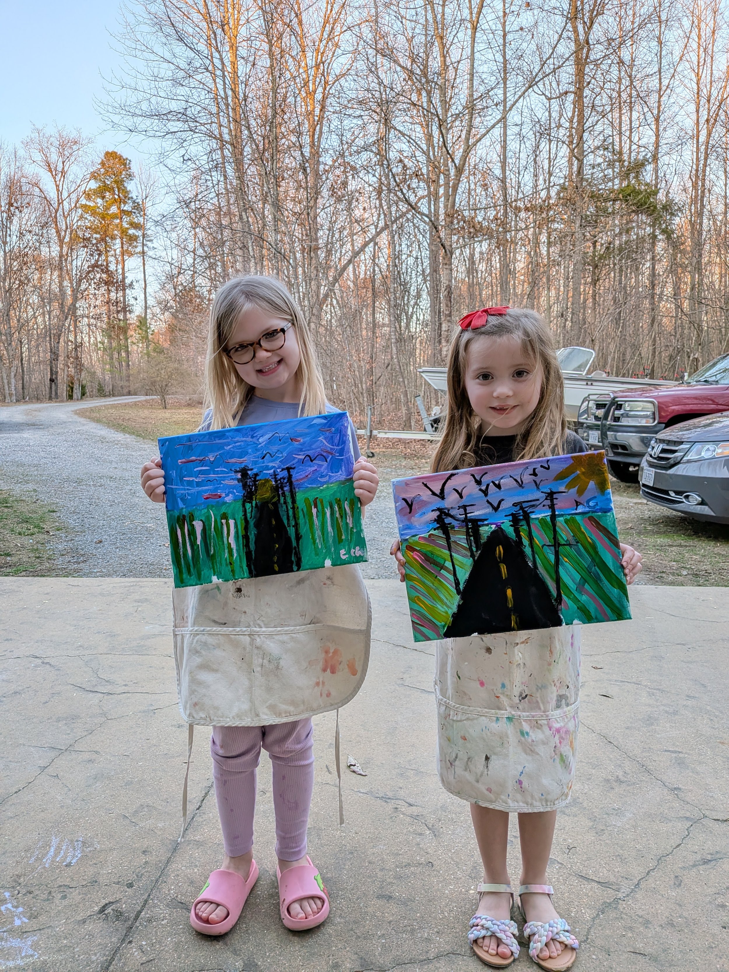 Two young girls standing outside on a concrete driveway, holding colorful paintings of a road with trees and birds in the sky. They are wearing aprons and smiling at the camera, with cars and trees in the background during sunset.