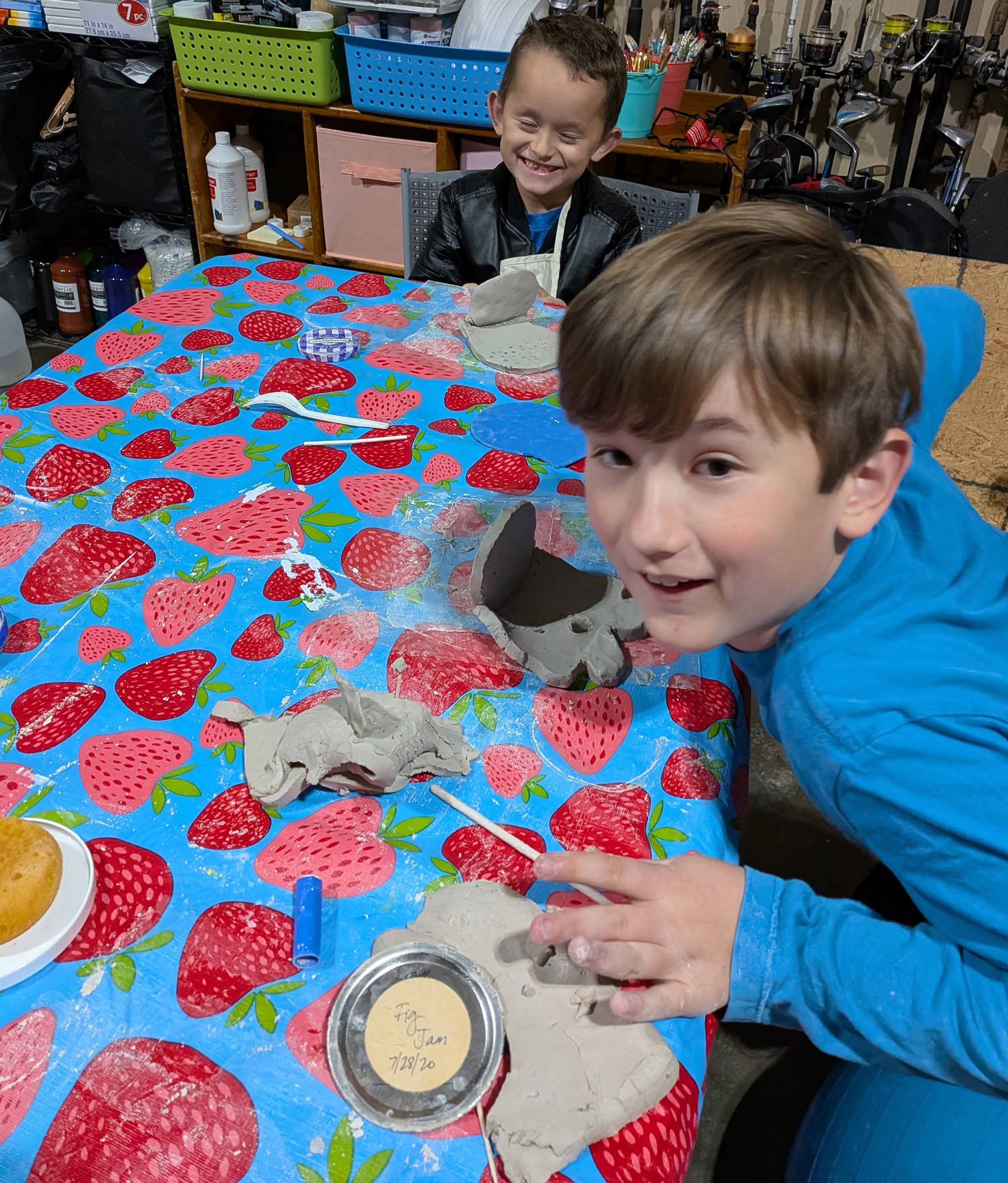 Two boys working on pottery projects at a craft table with a strawberry-themed tablecloth indoors, smiling and engaging in a creative activity.