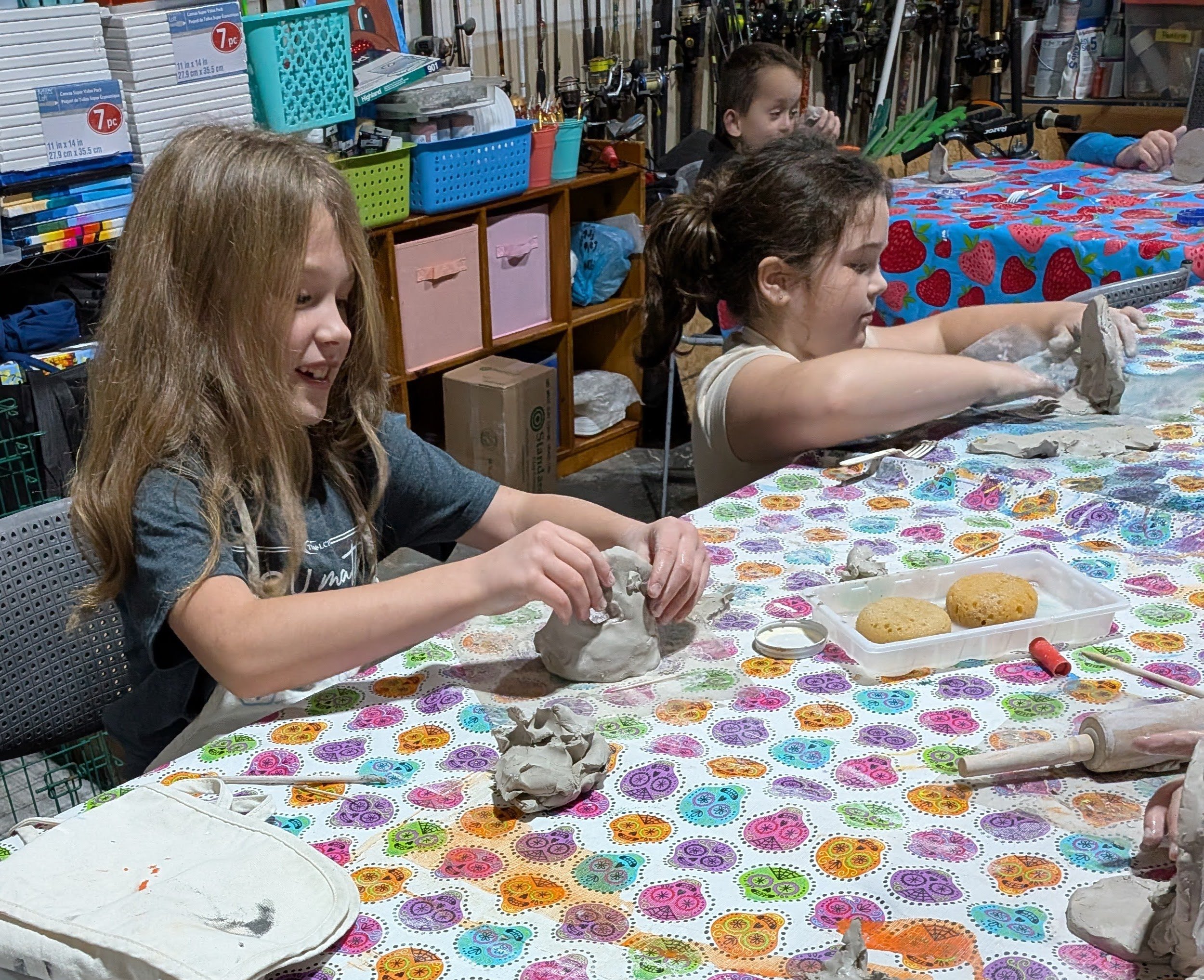 Children sitting at a colorful table, rolling and shaping clay, with art supplies and cupcakes nearby, in a craft workshop or classroom setting.