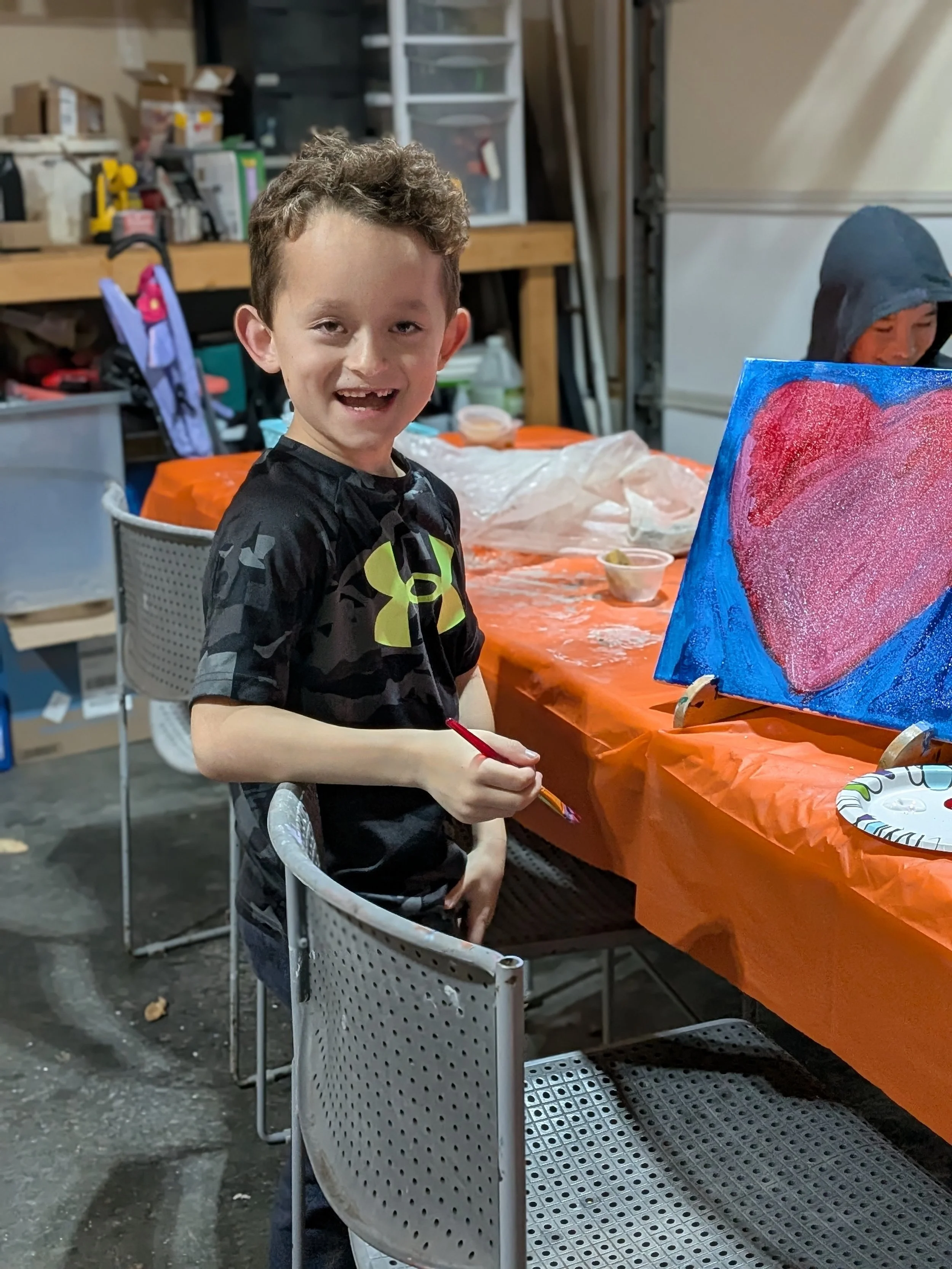 A young boy with short, curly hair smiling and holding a paintbrush while sitting at a table covered with an orange tablecloth. There is a painting of a pink heart on a blue background on the table, along with some small bowls of paint. The backgroun