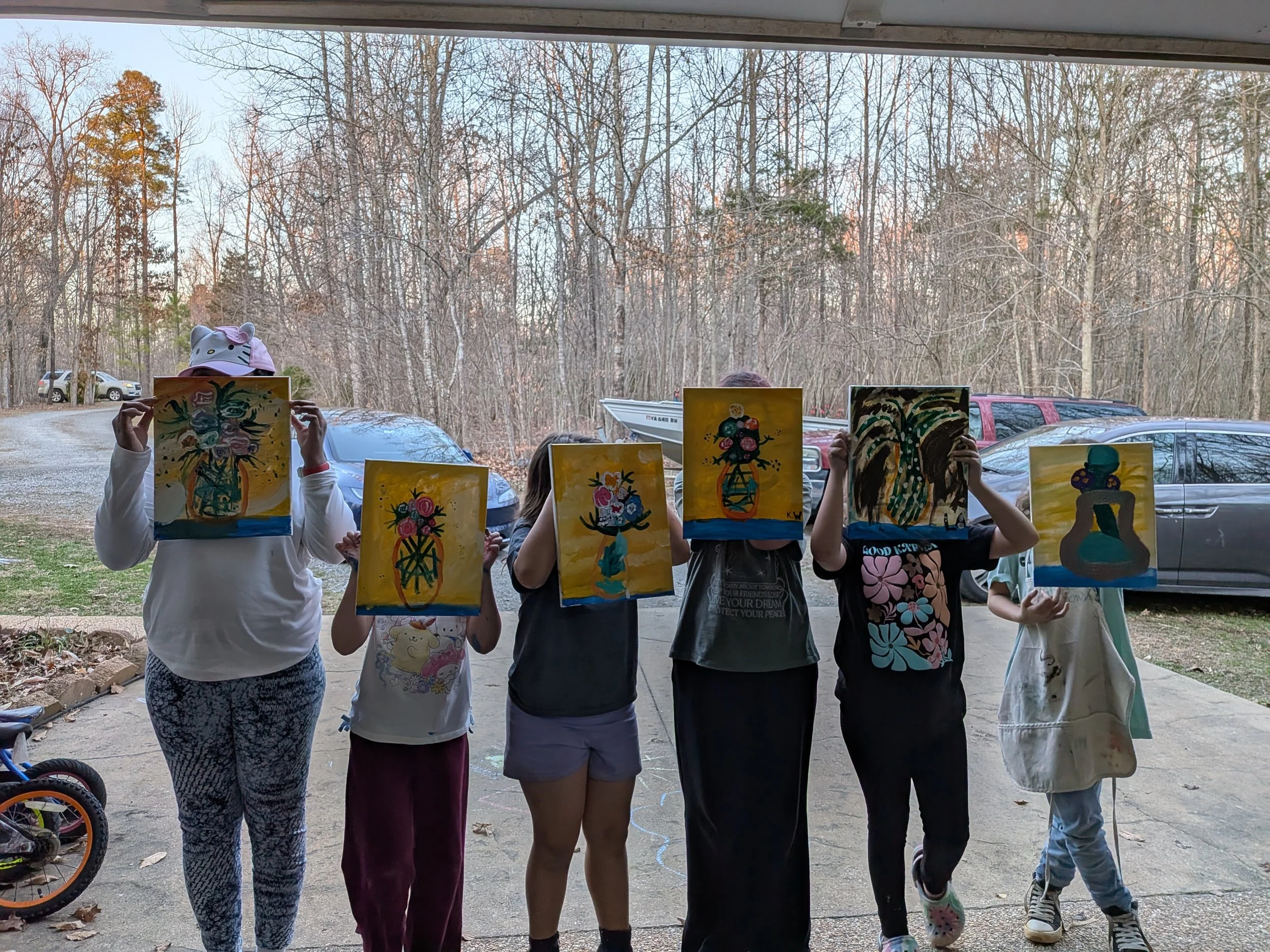 Six children standing outdoors in front of a wooded area, holding up paintings in front of their faces.