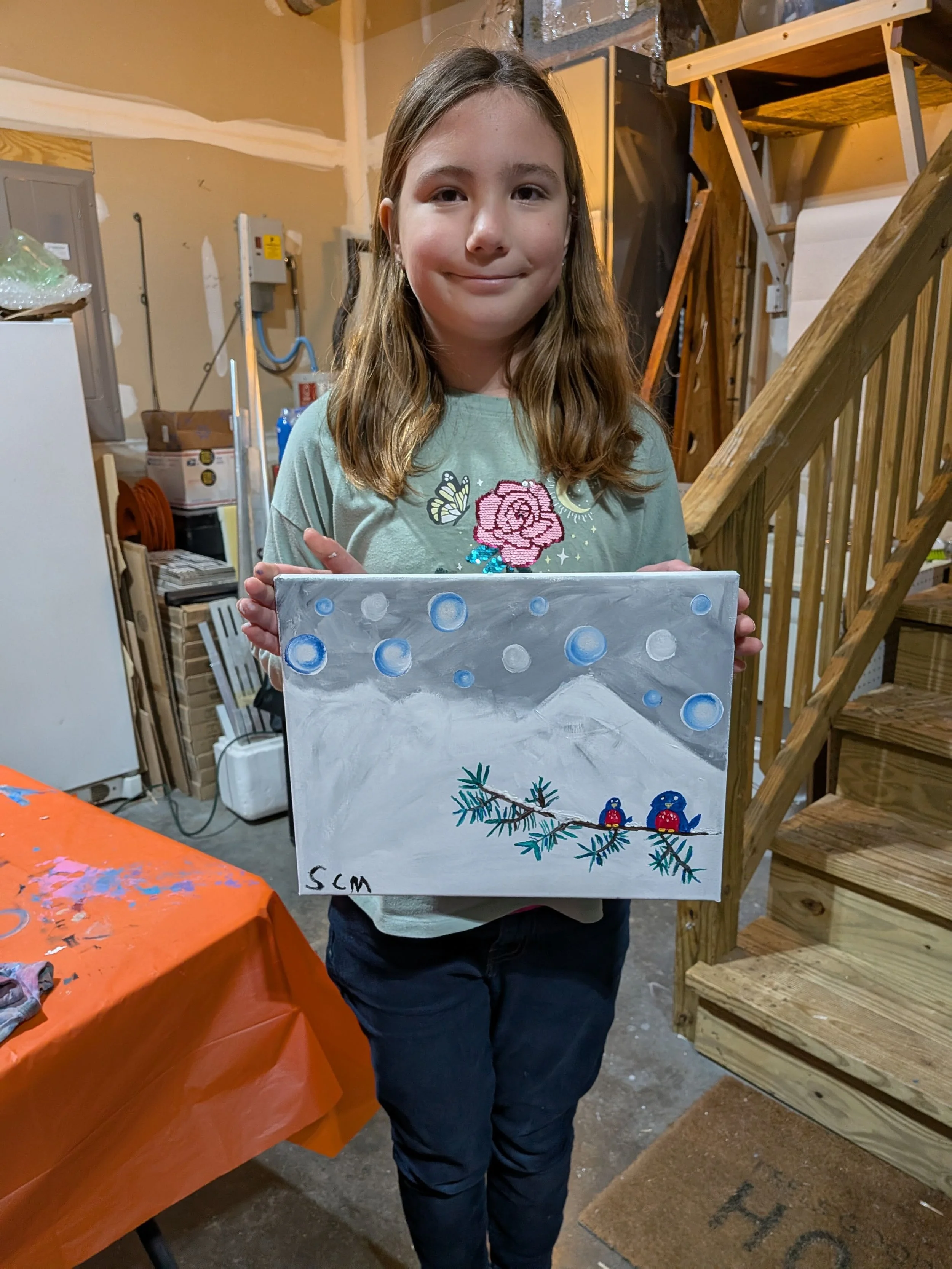 A young girl with shoulder-length hair holding a painting of a snowy mountain landscape with snow-covered trees and two birds sitting on a branch. She stands in a garage with wooden stairs and various tools and boxes in the background.