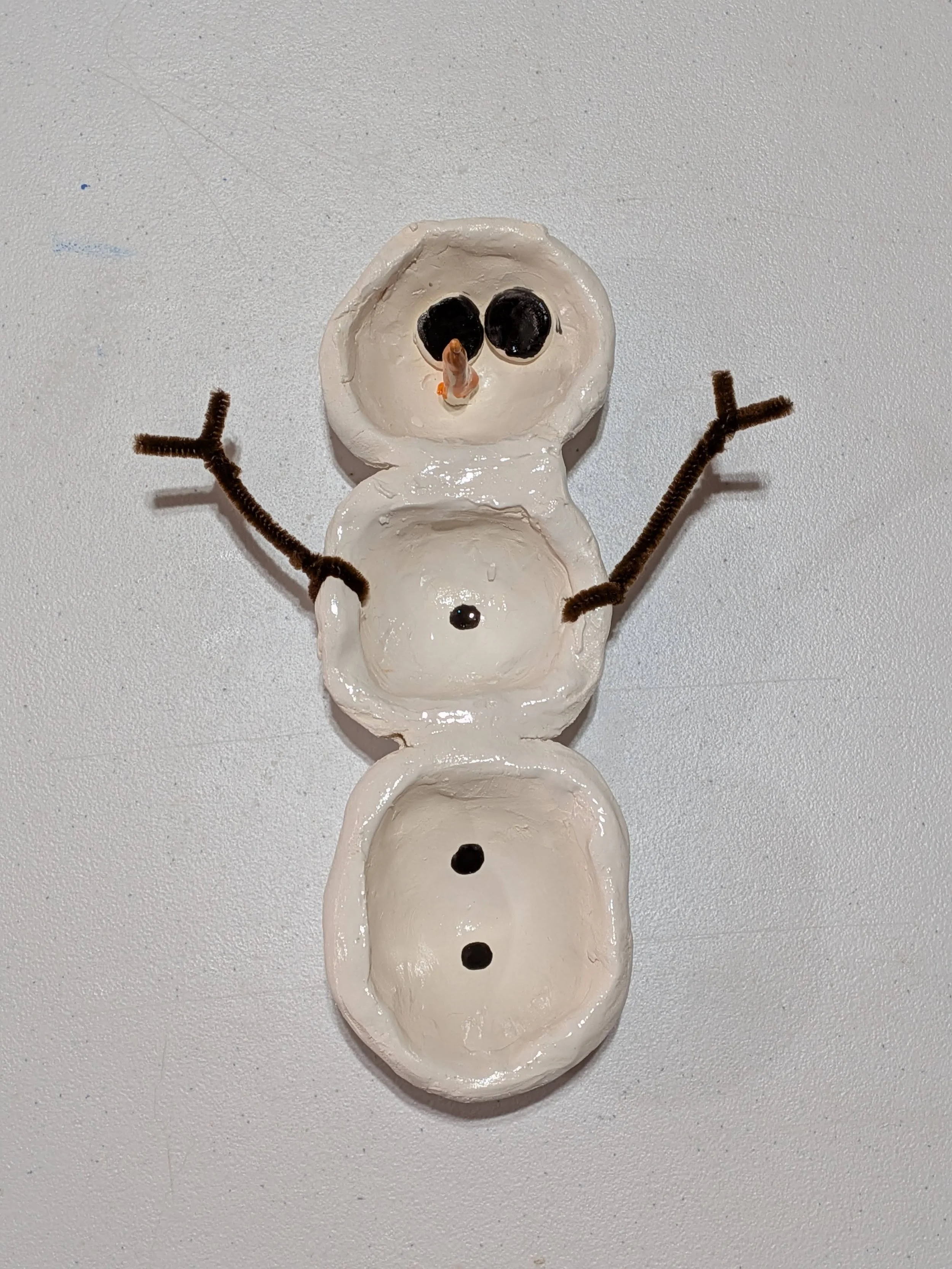 A snowman made from three empty egg shells, with black buttons and pipe cleaner arms, on a white surface.