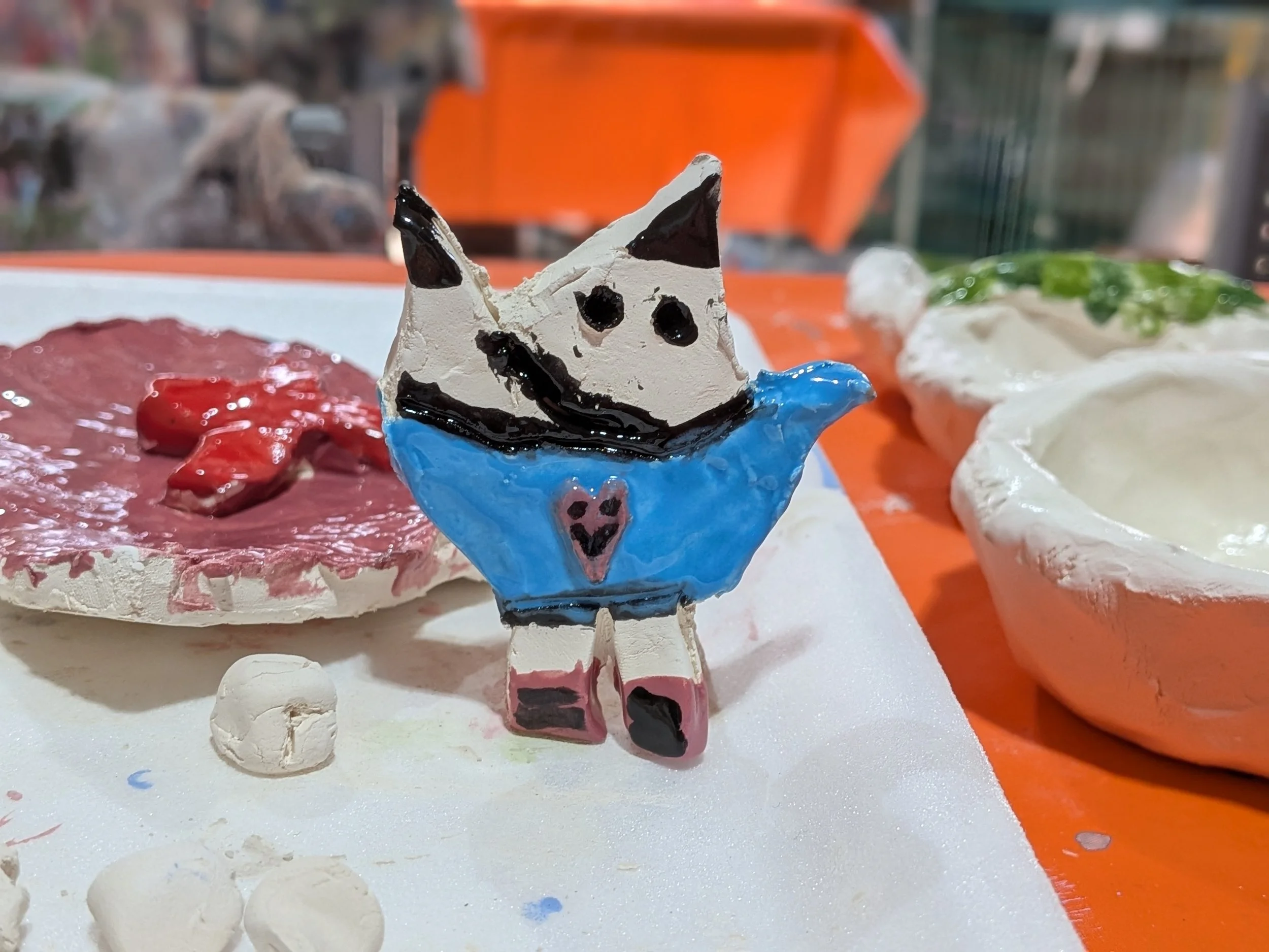 Colorful handmade ceramic figurine of a small animal, possibly a dog or cat, with a blue sweater and pink shoes, positioned next to painted ceramic pottery on a work surface.