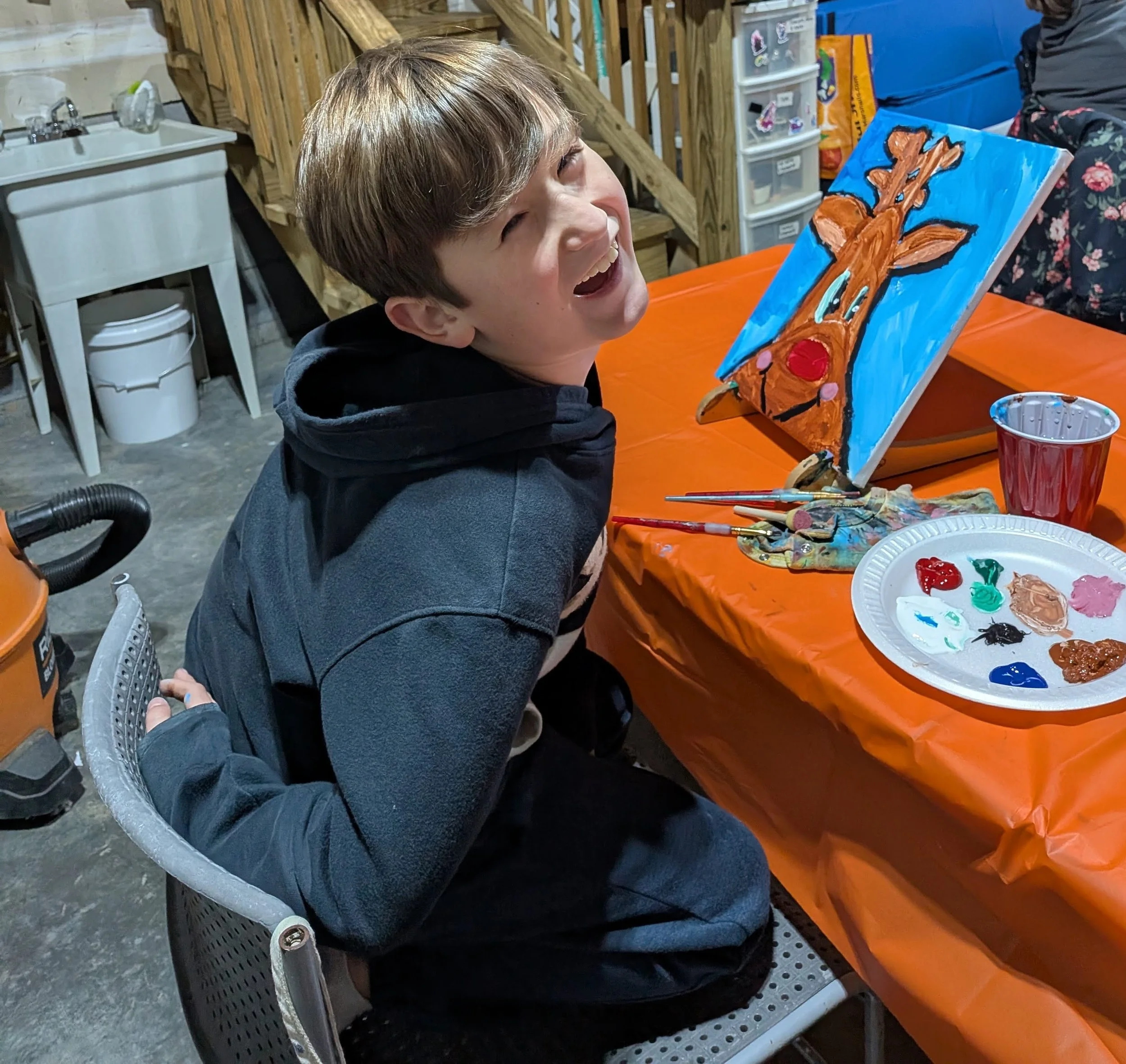 A boy sitting at a table with a completed painting of a reindeer's head, painted with brown and red colors on a blue background. The table has a palette with various paint colors, paintbrushes, and a red cup.