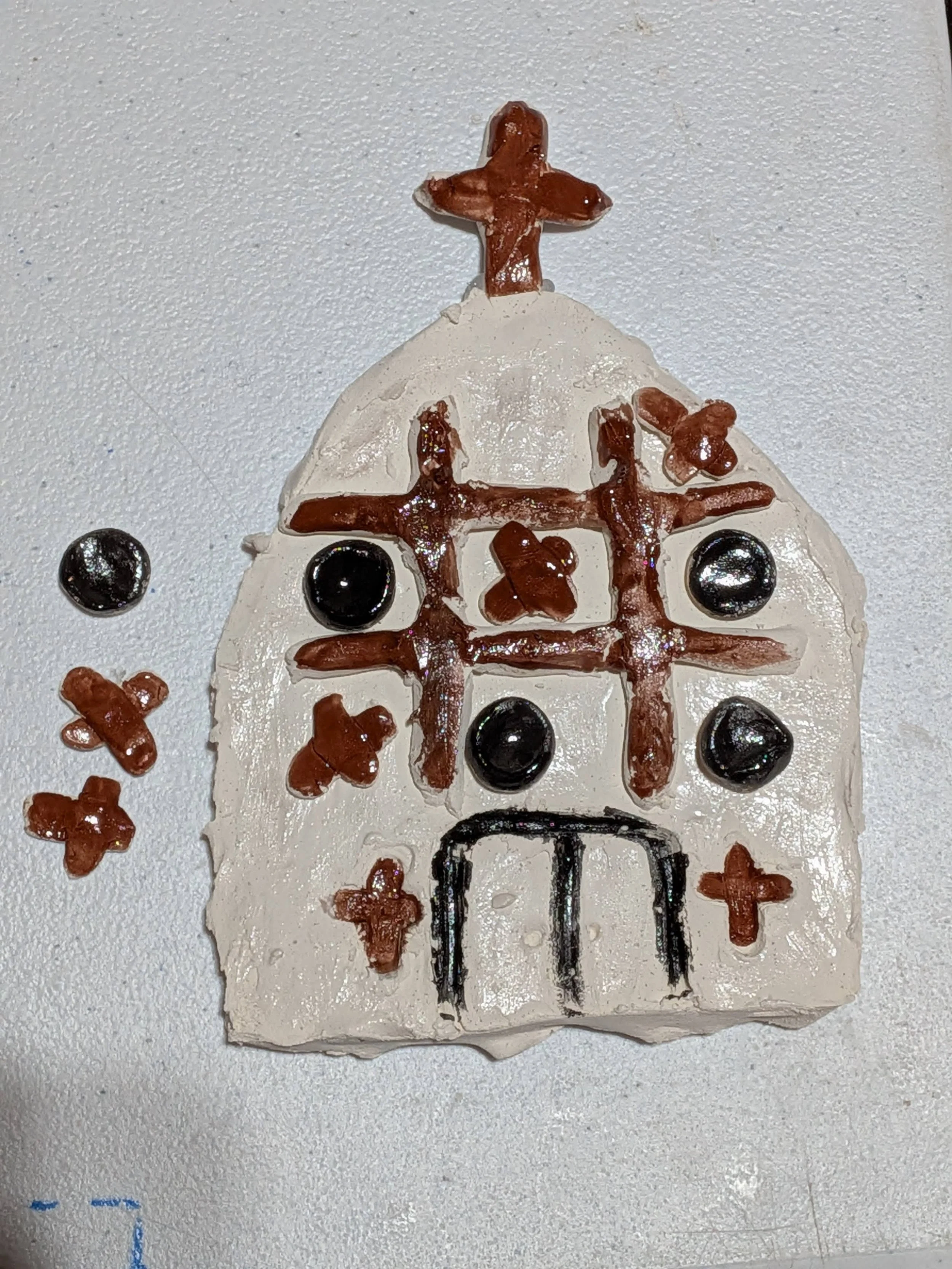 A craft representation of a church made from white and brown materials, with black dots for windows and a simple black door. The church has a cross at the top and decorative crosses on the walls.