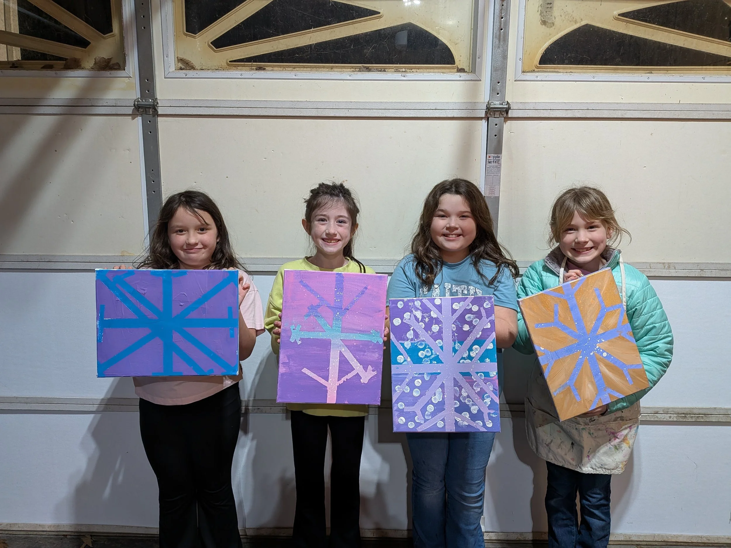 Four girls proudly holding painted snowflake designs on canvases in front of a garage door