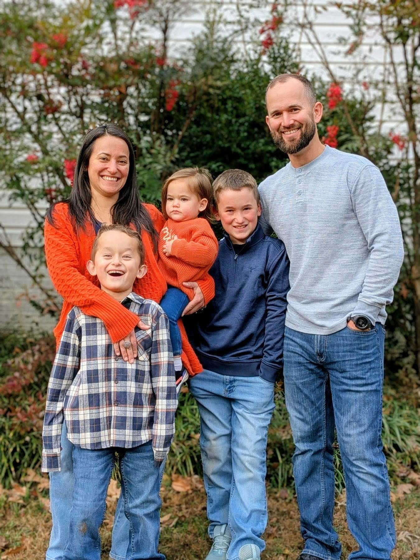 Family of five outdoors smiling, with a house and bushes with red flowers in the background.