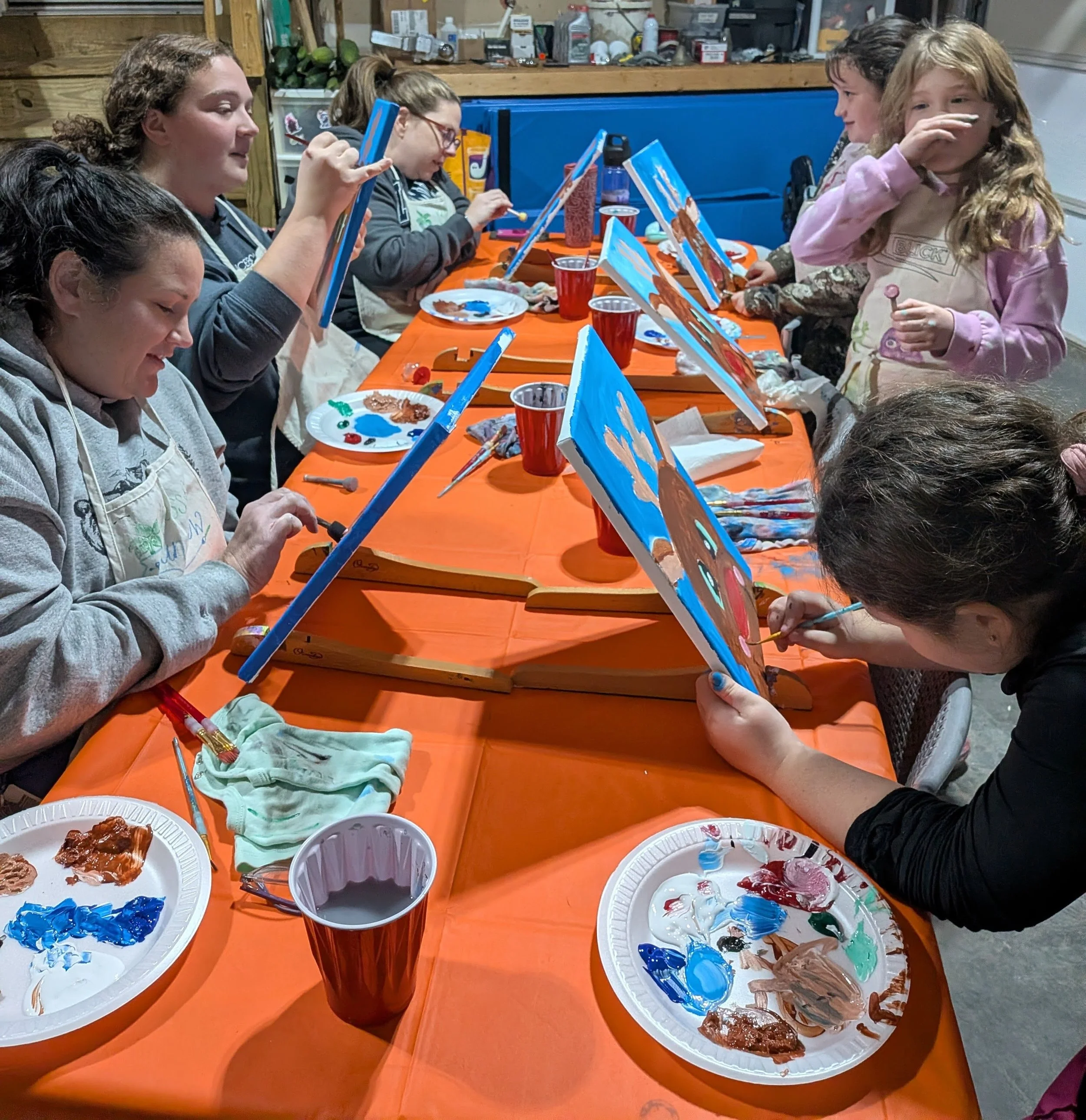 Children participating in a group painting activity at a long table with orange tablecloths, paint palettes, and red cups filled with water, in a room with shelves and cabinets in the background.