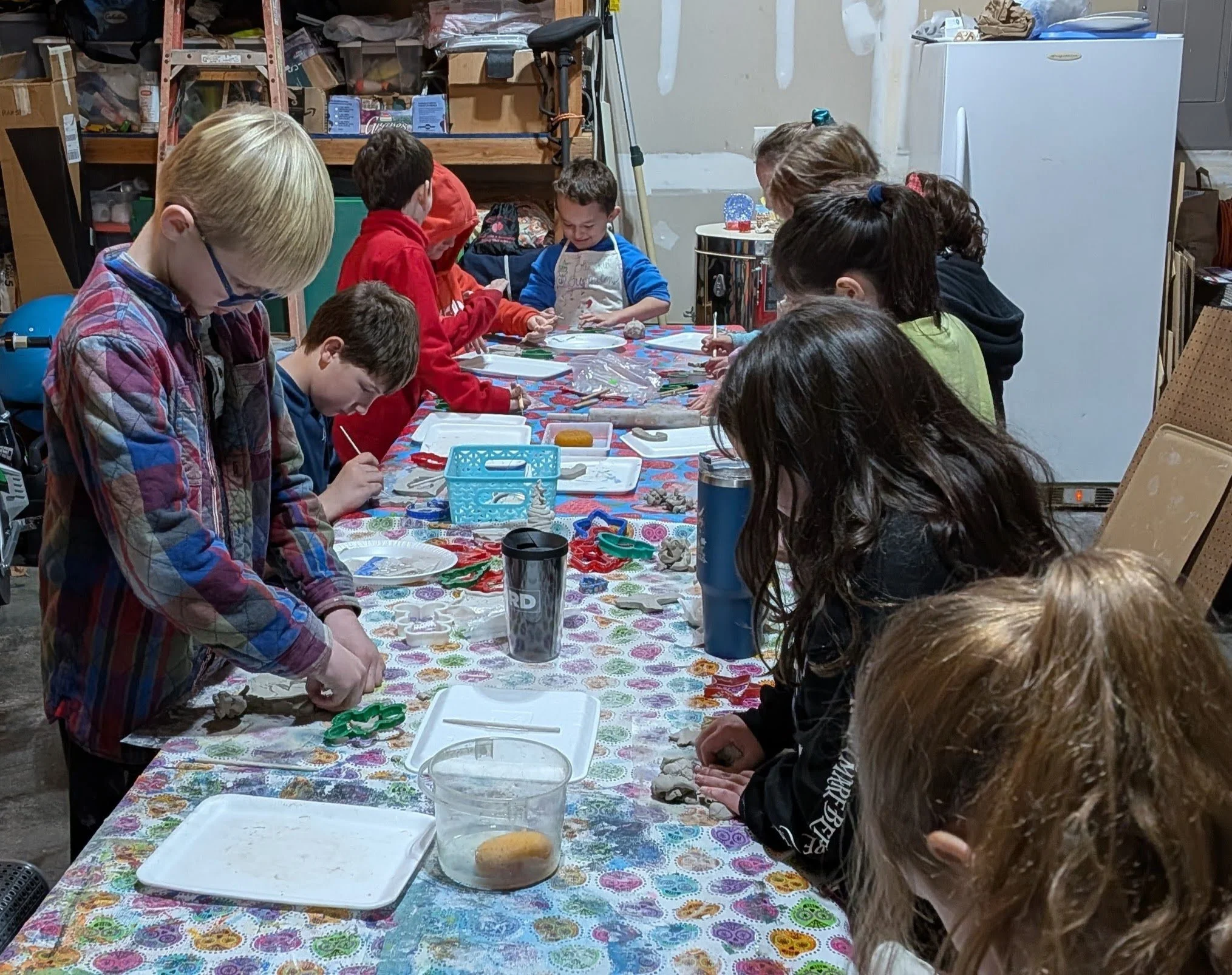 Children participating in a craft activity around a long table covered with a colorful tablecloth in a garage or workshop setting.