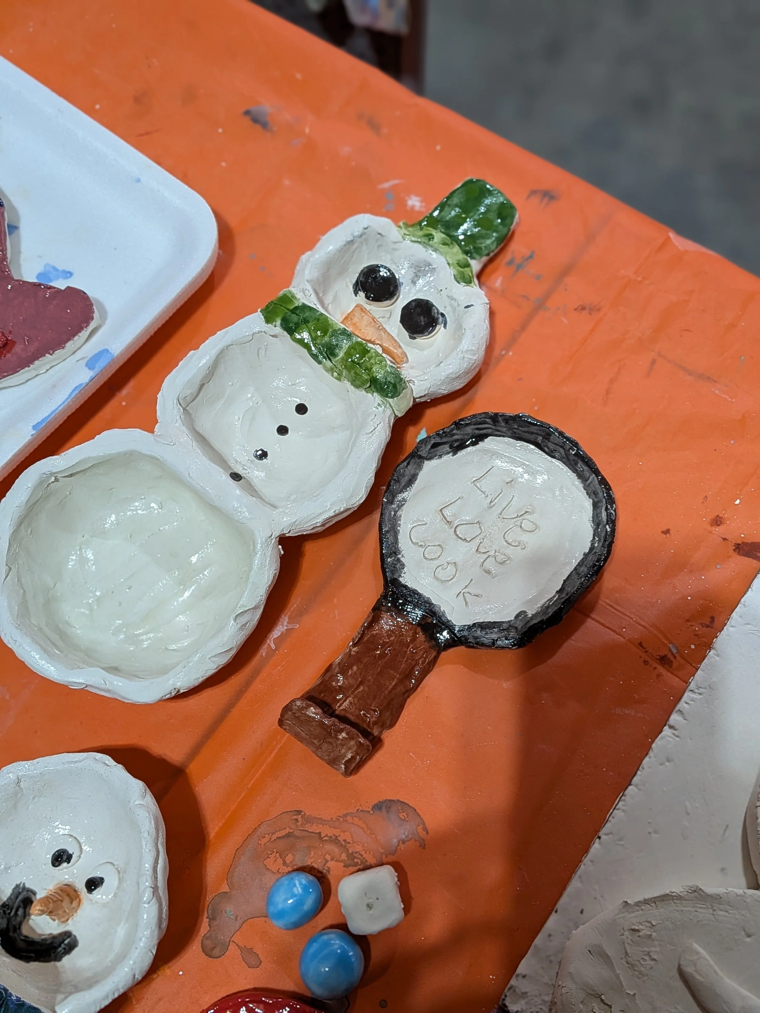 Snowman craft made from painted shells with leaves for hat and scarf, black buttons, and googly eyes, along with a small wooden-handled mirror that reads 'I love you' and small painted rocks.