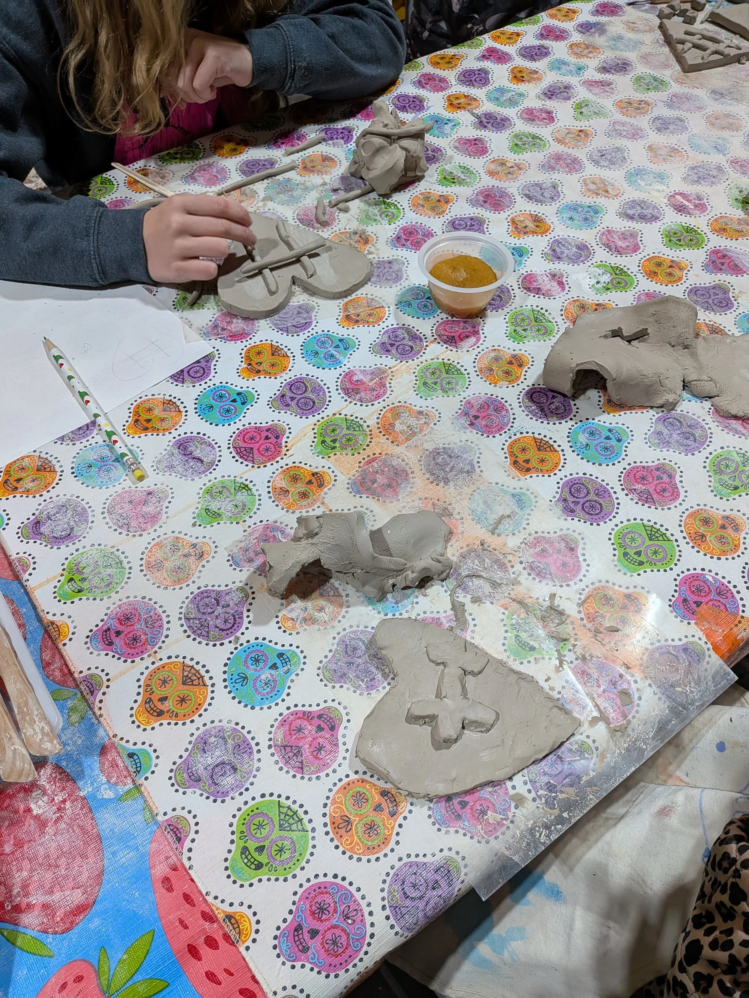 Children working on clay sculptures at a decorated table with Day of the Dead themed paper tablecloths.