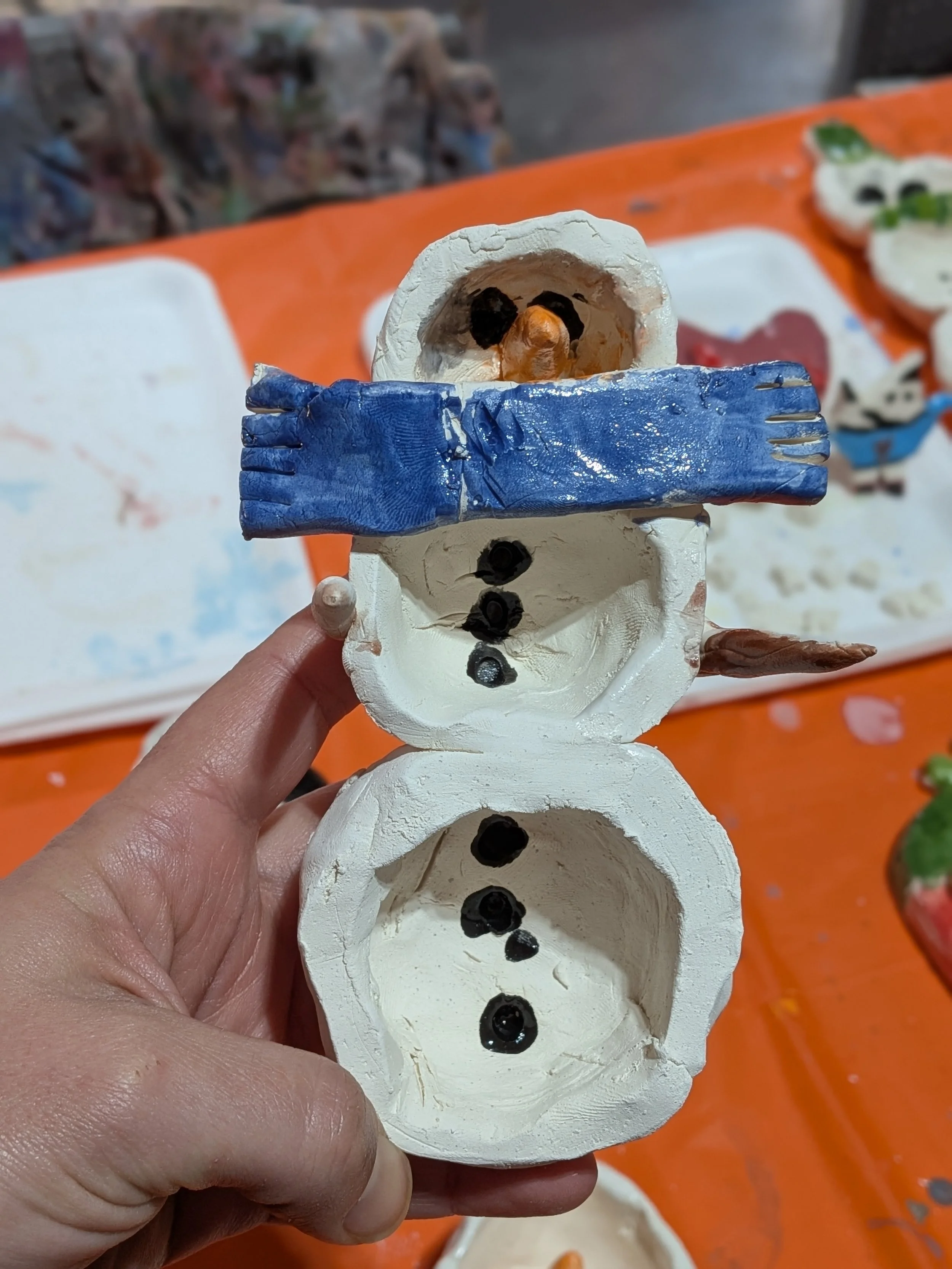 A handmade snowman craft made from painted paper or cardboard, with a face, blue scarf, and black buttons, held in a person's hand, with other painted crafts visible on a table in the background.