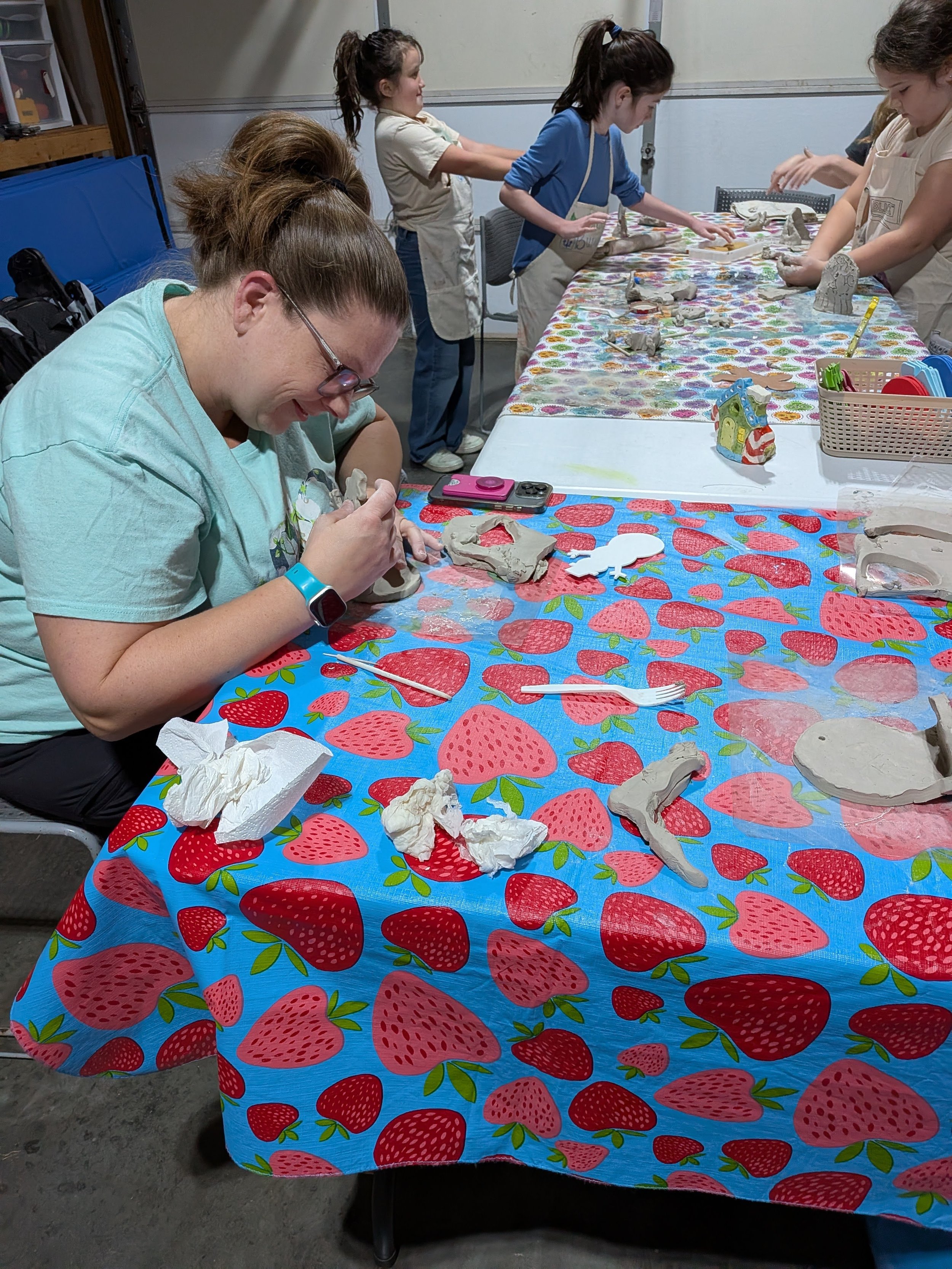 People participating in a ceramic painting activity at a table covered with a strawberry-patterned tablecloth. One woman in the foreground is focused on her ceramic piece, with others working in the background.
