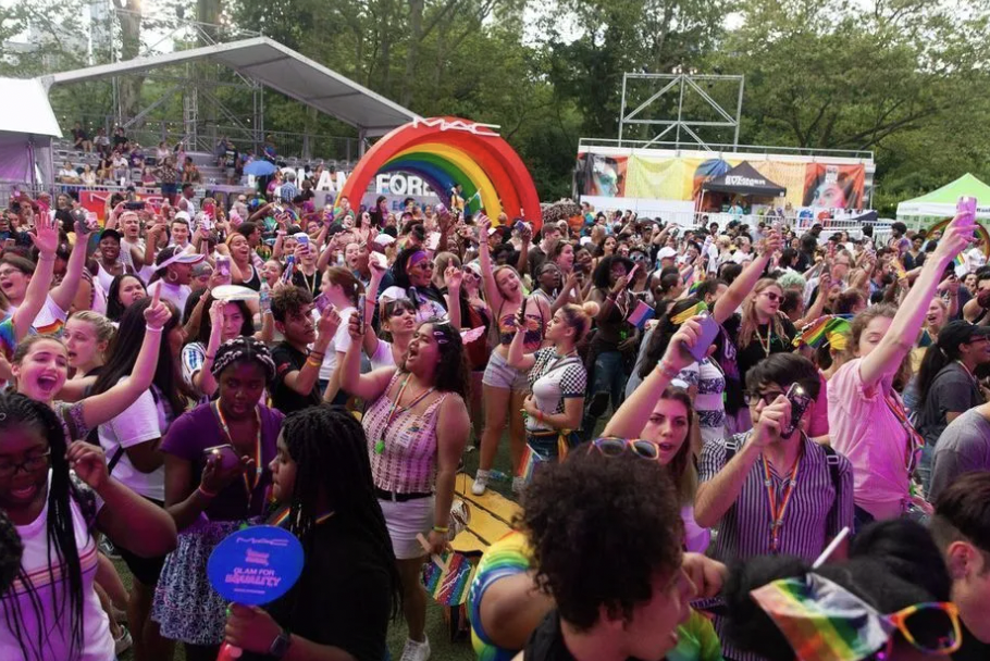 Large crowd of people at a pride event outdoors with rainbow-themed decorations, including a rainbow rainbow-shaped arch and rainbow flags, celebrating LGBTQ+ pride.