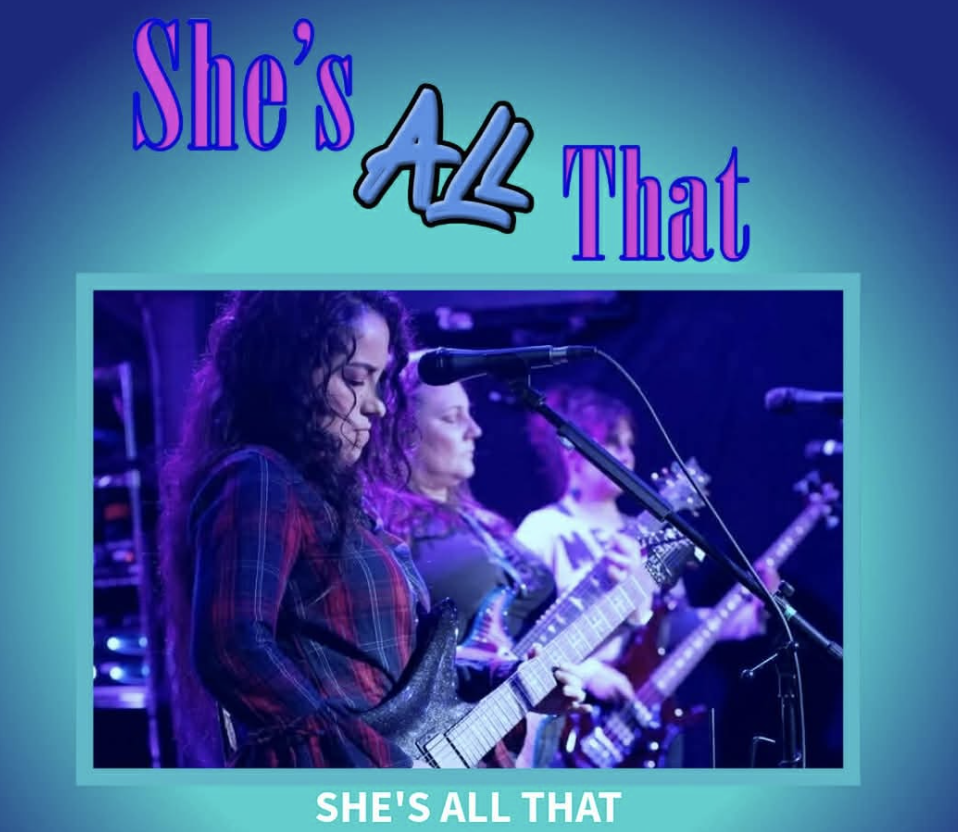 A group of women musicians performing live on stage with a sign behind them that reads "The Femmes at Silver Lining Lounge." The women are singing and playing instruments, with some raising their arms in celebration. The scene is vibrant with stage lighting and a disco ball overhead.