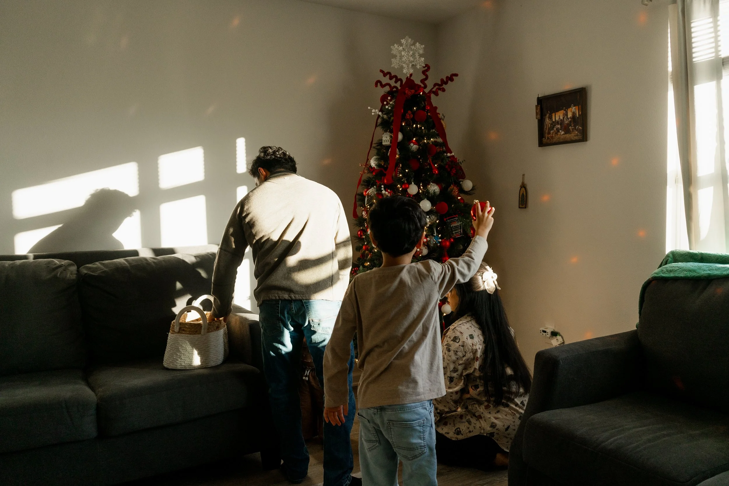 People decorating a Christmas tree in a living room, with sunlight casting shadows through window blinds.