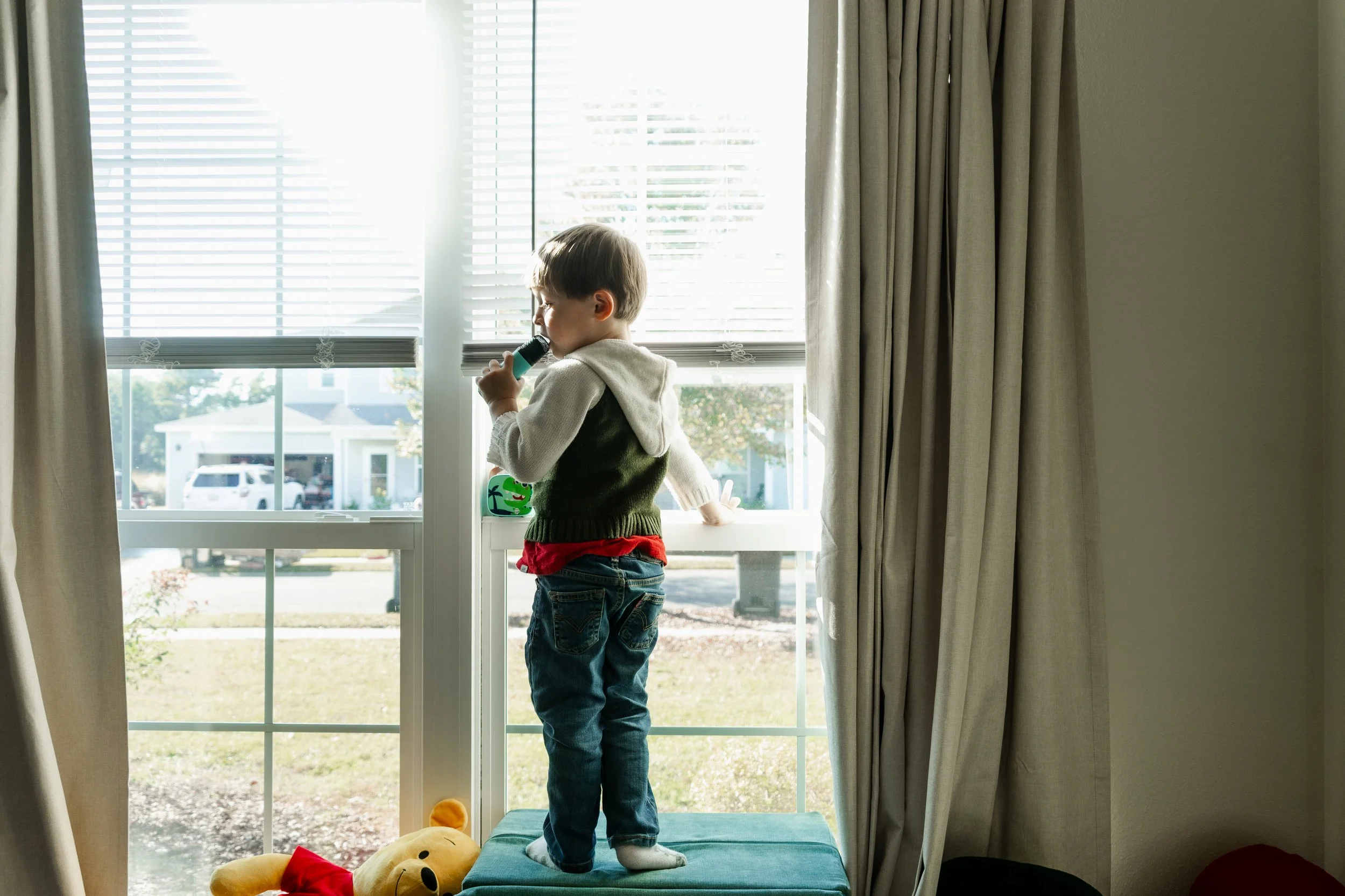 A young boy standing on a window seat, looking out and holding a toy microphone, with a teddy bear on the floor nearby.