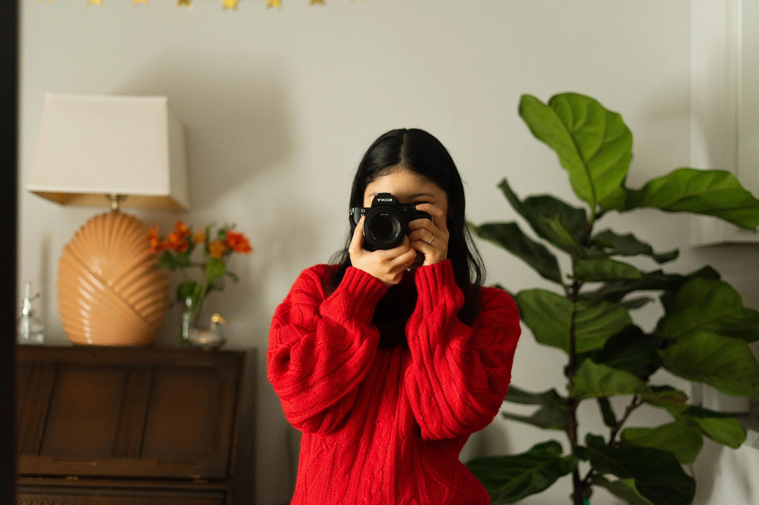 Woman in red sweater taking a selfie in front of a mirror, with a large green houseplant, a wooden cabinet, a lamp, and a vase with orange flowers in the background.
