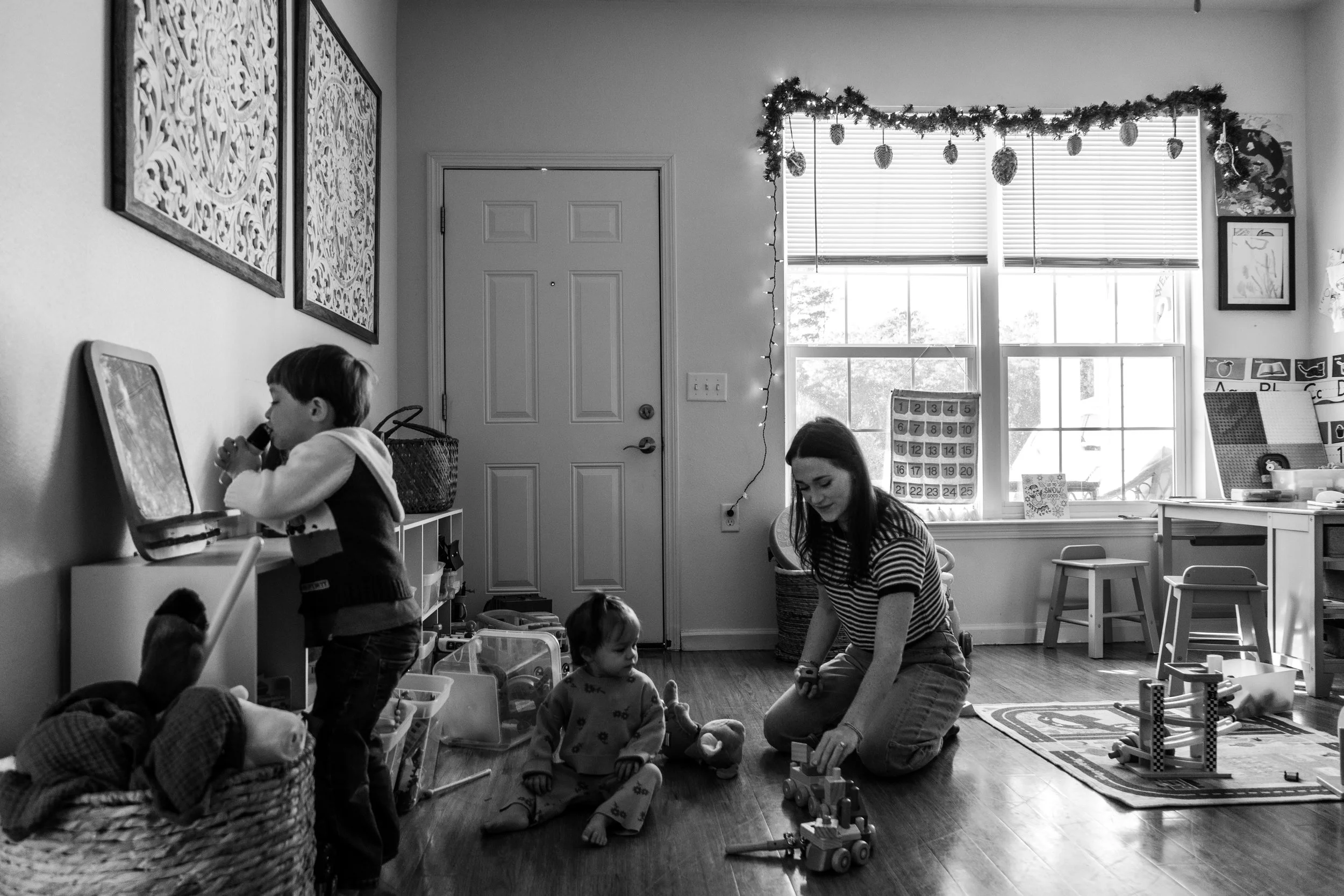 A woman playing with two young children on a hardwood floor in a playroom decorated with holiday garland. One child is standing and drinking from a cup, while the other is sitting with toys. The room has windows, decorations, and storage furniture.