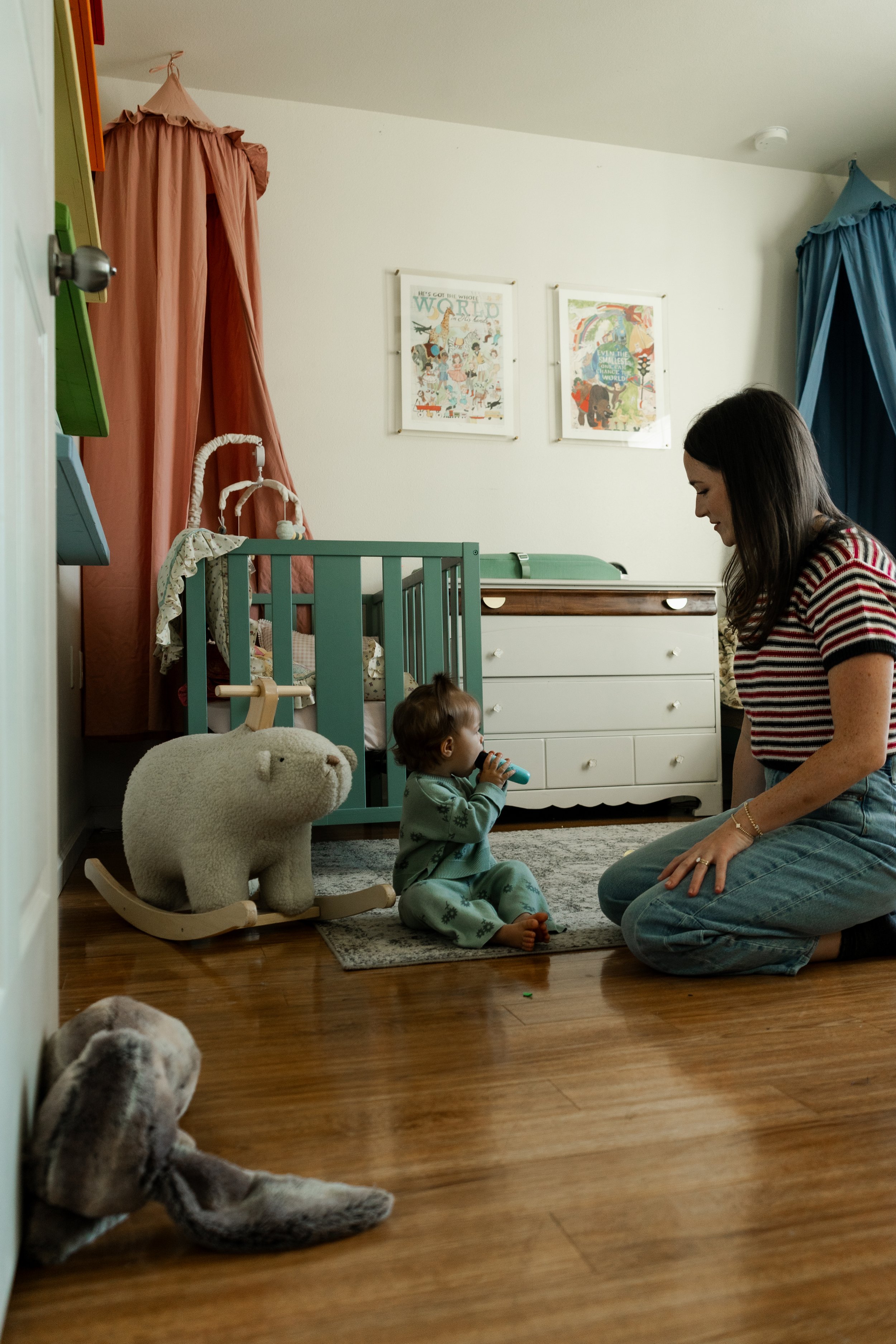 A woman kneeling on the floor next to a small child sitting on a rug in a nursery room, with toys including a plush bear on a rocking base nearby.