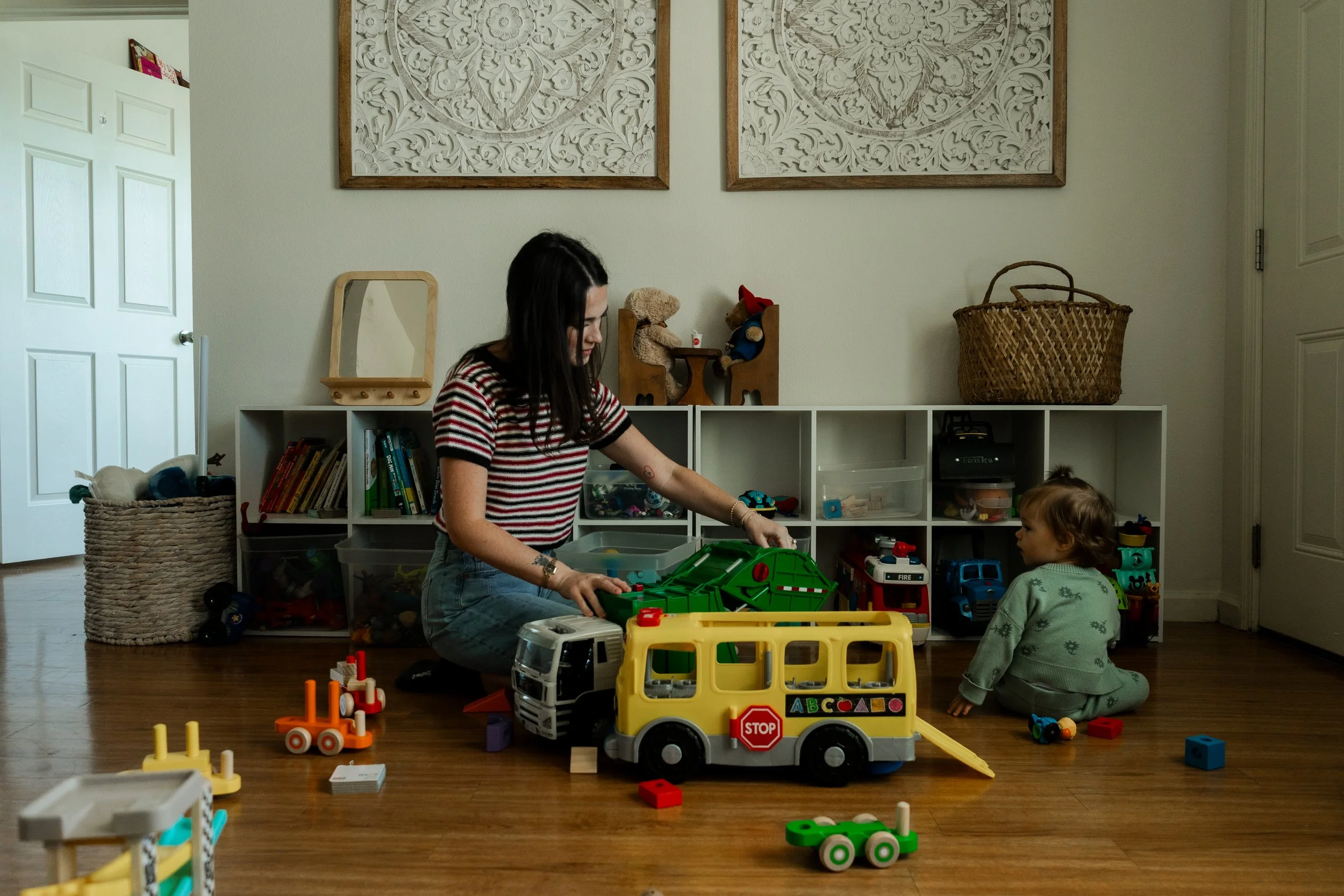 A woman and a small child playing on the floor with colorful toy vehicles and blocks in a living room. The woman is sitting, and the child is kneeling, surrounded by toy buses and cars. Behind them, a white storage unit with toys and books, framed wall art, and a basket on top are visible.