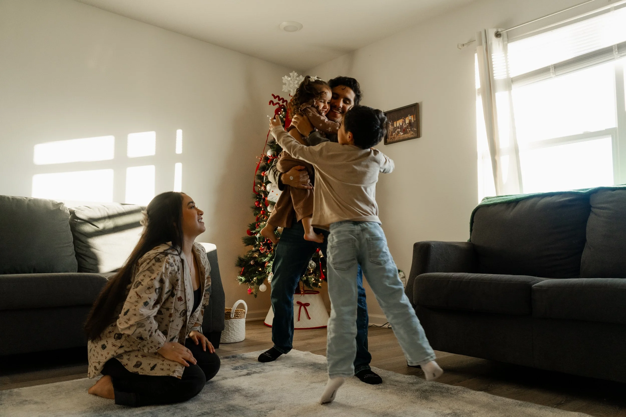 A family celebrating Christmas in a living room with a decorated Christmas tree. A man holding a young girl is being lifted by a boy, while a woman kneels and smiles nearby.