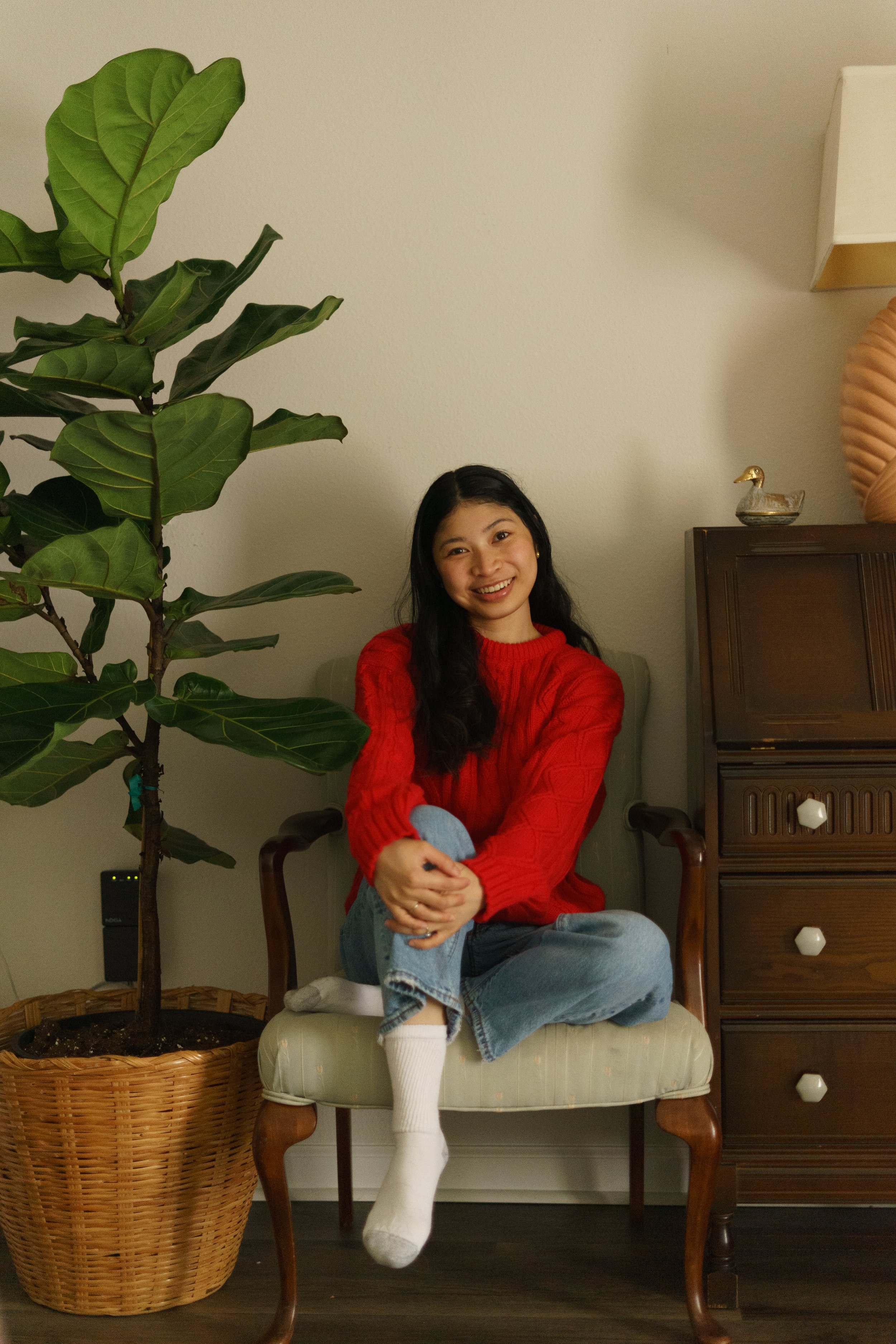 A woman sits on a vintage armchair with a light-colored cushion, wearing a red sweater, blue jeans, and white socks, smiling at the camera. To her left is a tall potted plant with large green leaves, and to her right is a wooden chest of drawers with decorative knobs and a ceramic duck figurine on top.
