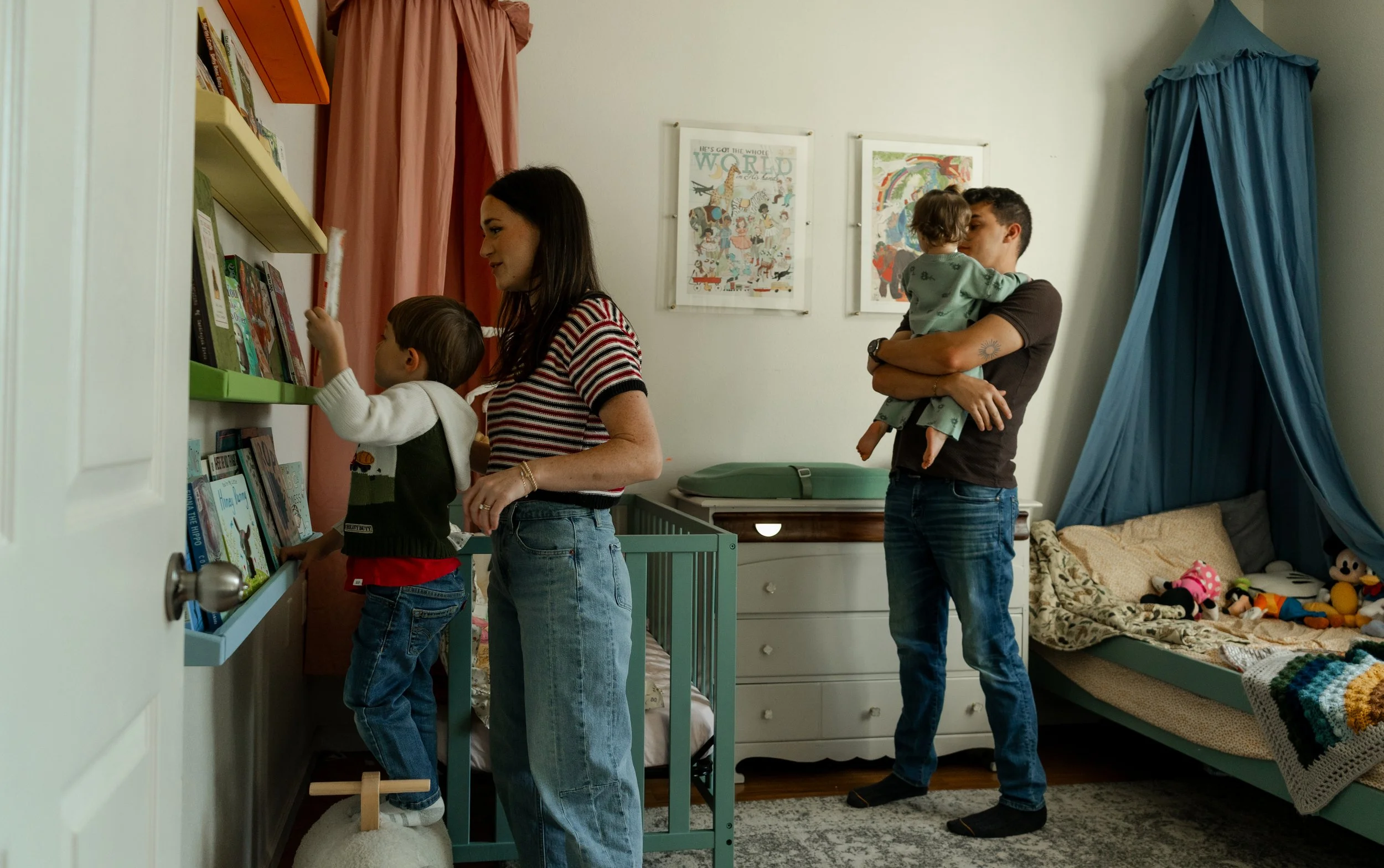 A woman and two children in a bedroom, with one child reaching for books on a shelf and the woman looking on, while the man holds a young child in front of another wall decorated with framed pictures.