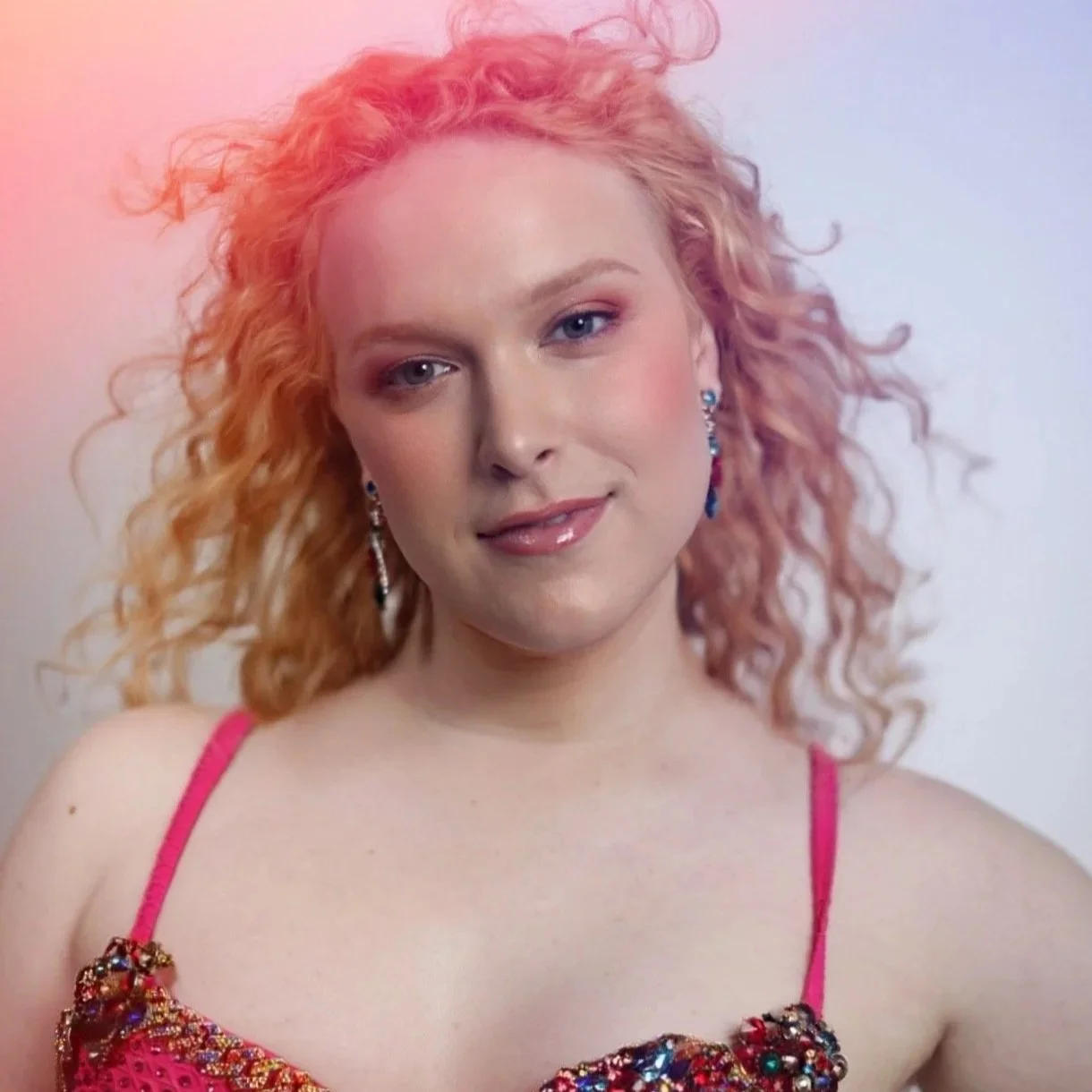 A young woman with curly pink hair and fair skin, wearing pink lipstick, eye makeup, and blue earrings, dressed in a sparkly pink top with thin straps.