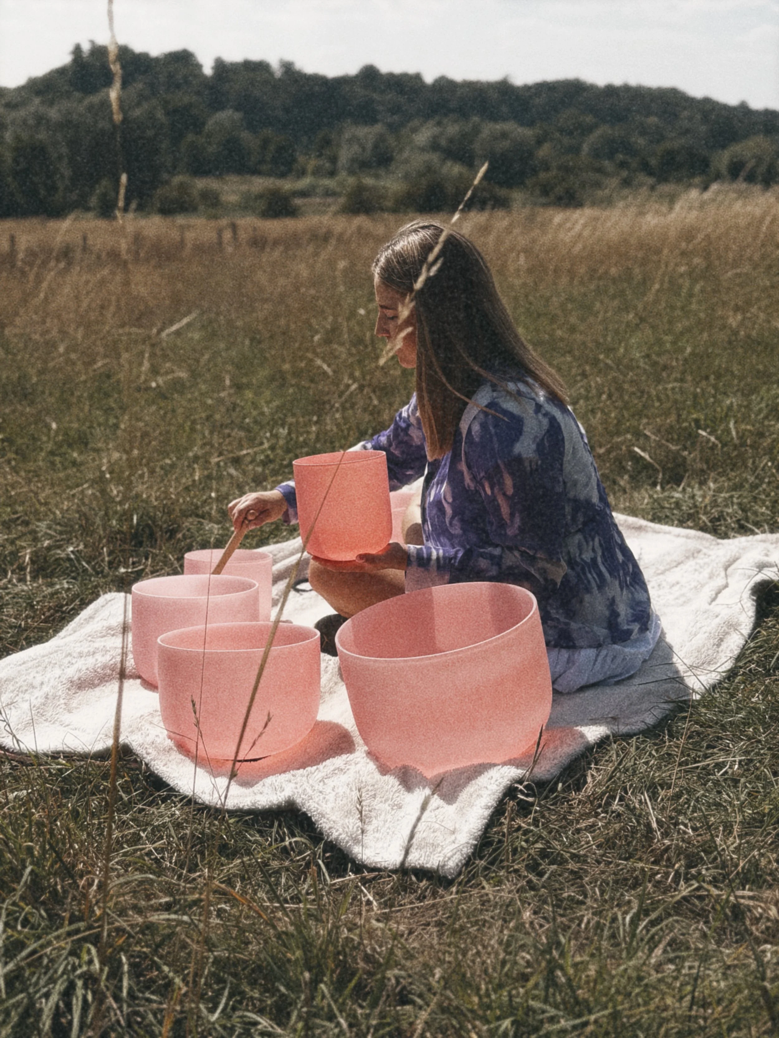 A woman is sitting on a white towel in a grassy field, playing crystal singing bowls. The landscape has rolling hills and trees in the background under a partly cloudy sky.