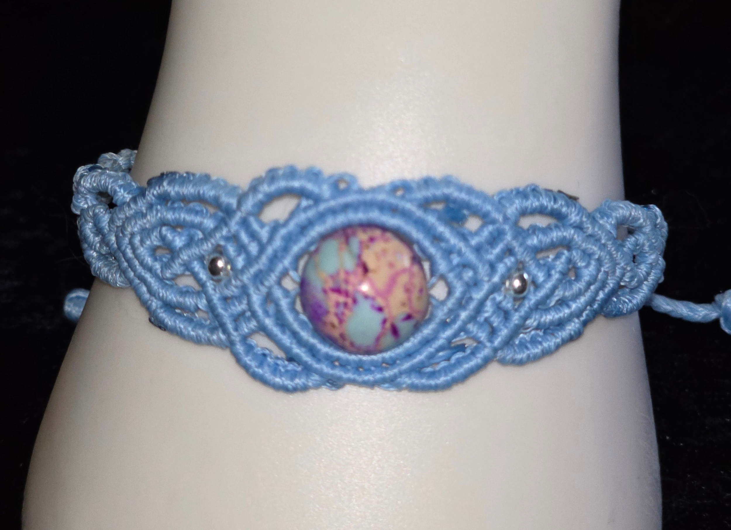 Light blue woven bracelet with a large marbled bead at the center, small metal beads on each side, and a black background.