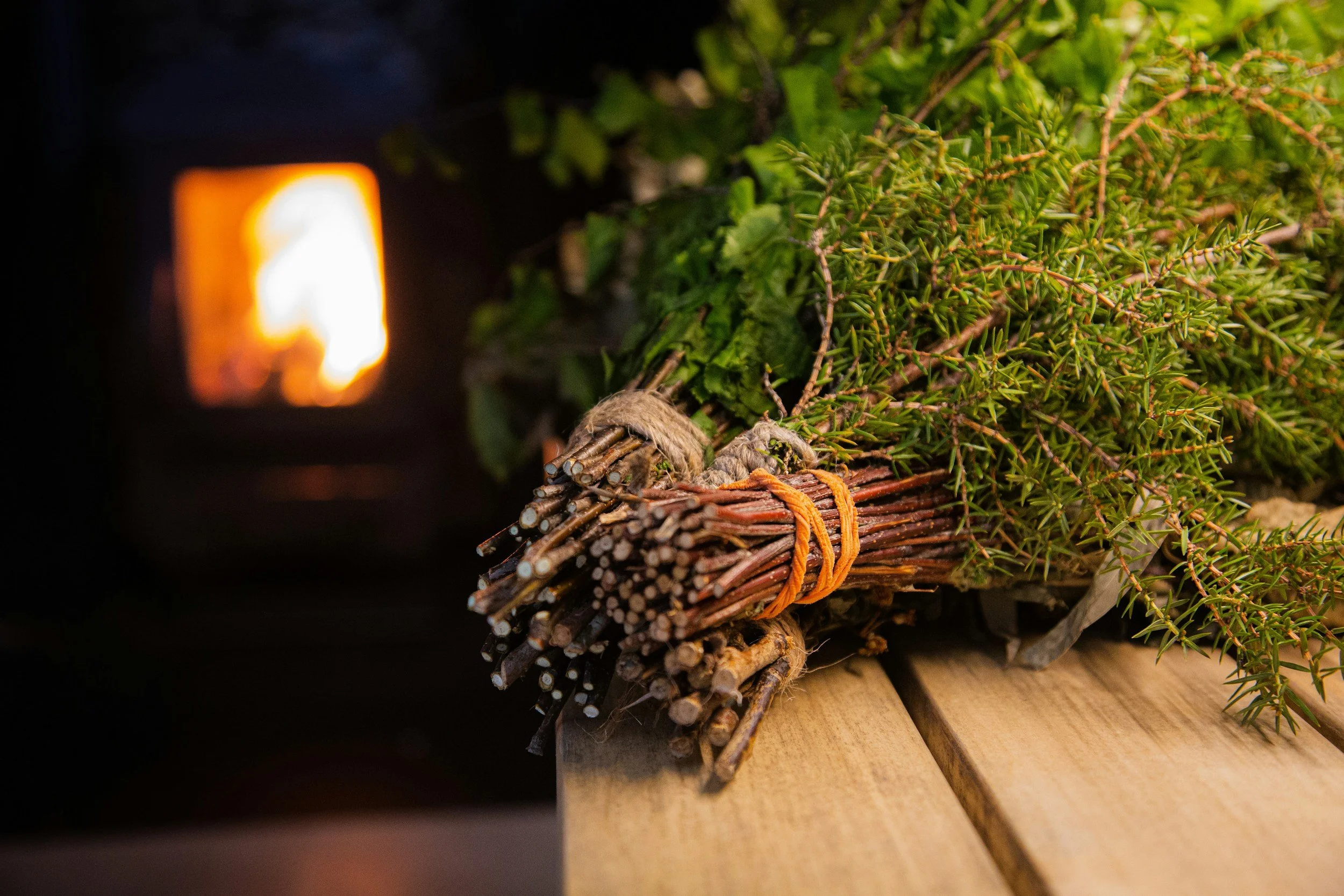 A bundle of small sticks tied with orange string, with green foliage on top, resting on a wooden surface near a fireplace.