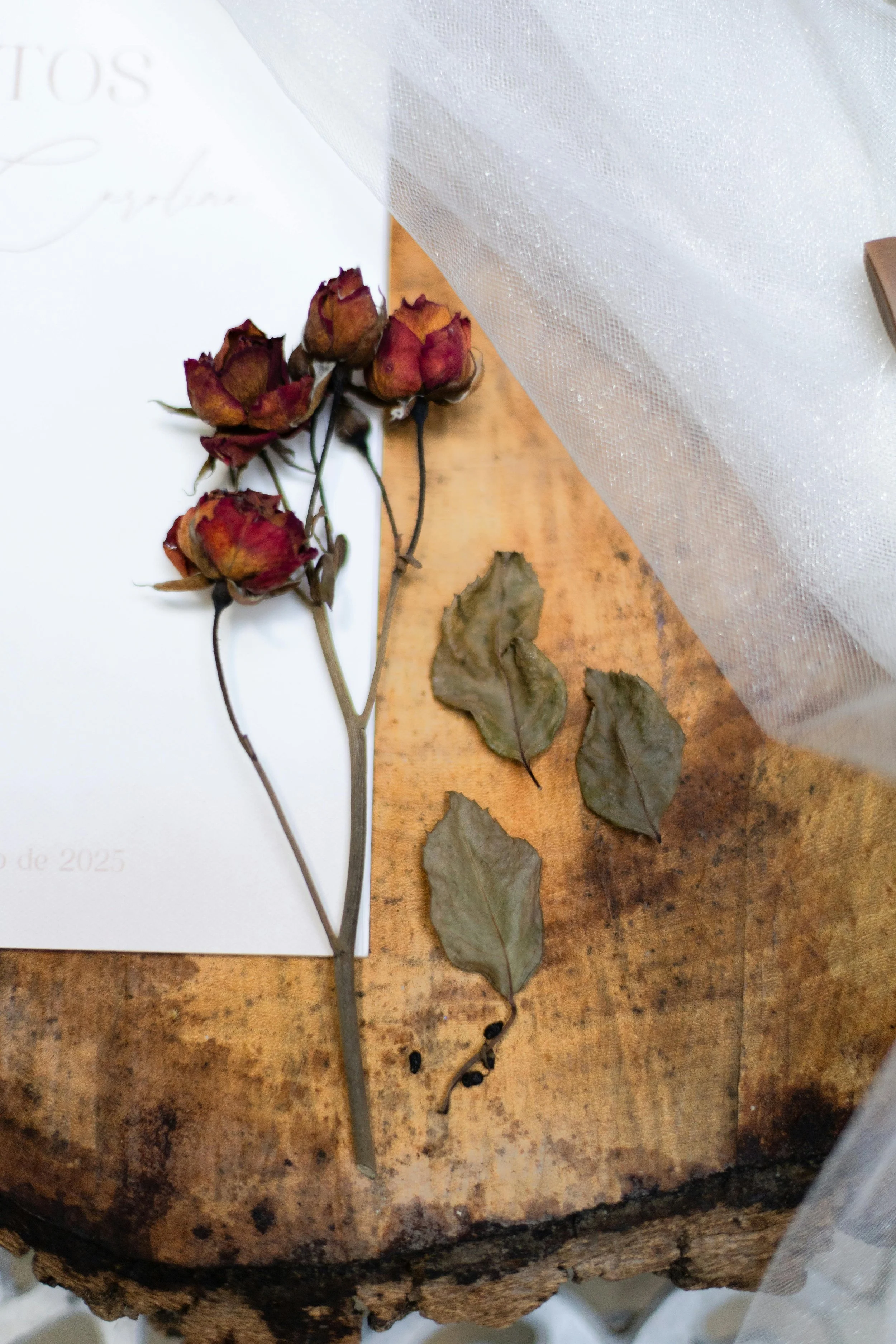 Dried red roses, dried leaves, and black peppercorns on a wooden surface beside a white piece of paper.