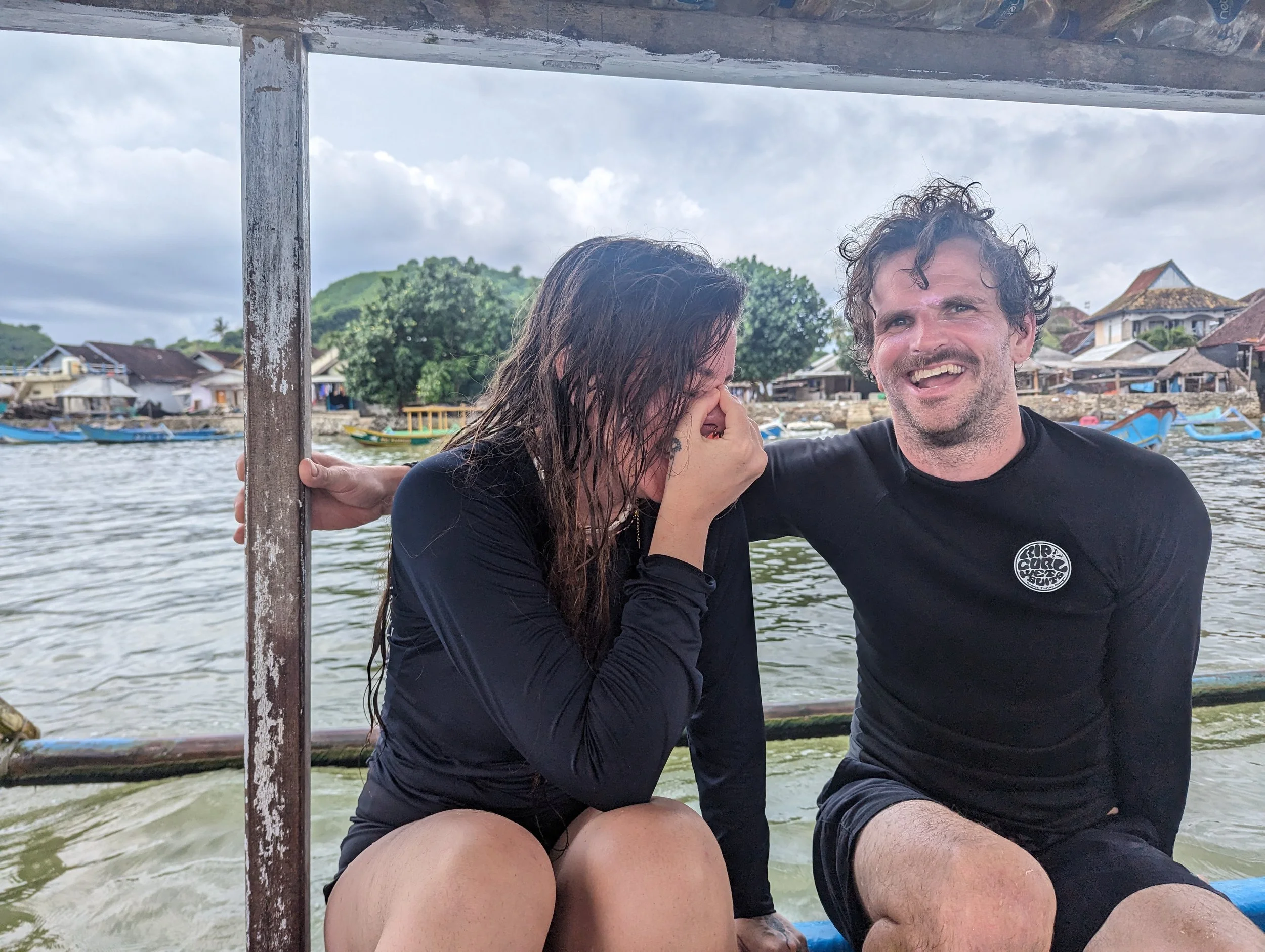 A couple sitting on a boat, smiling and laughing during a rainy day near a riverside village with boats and houses in the background.