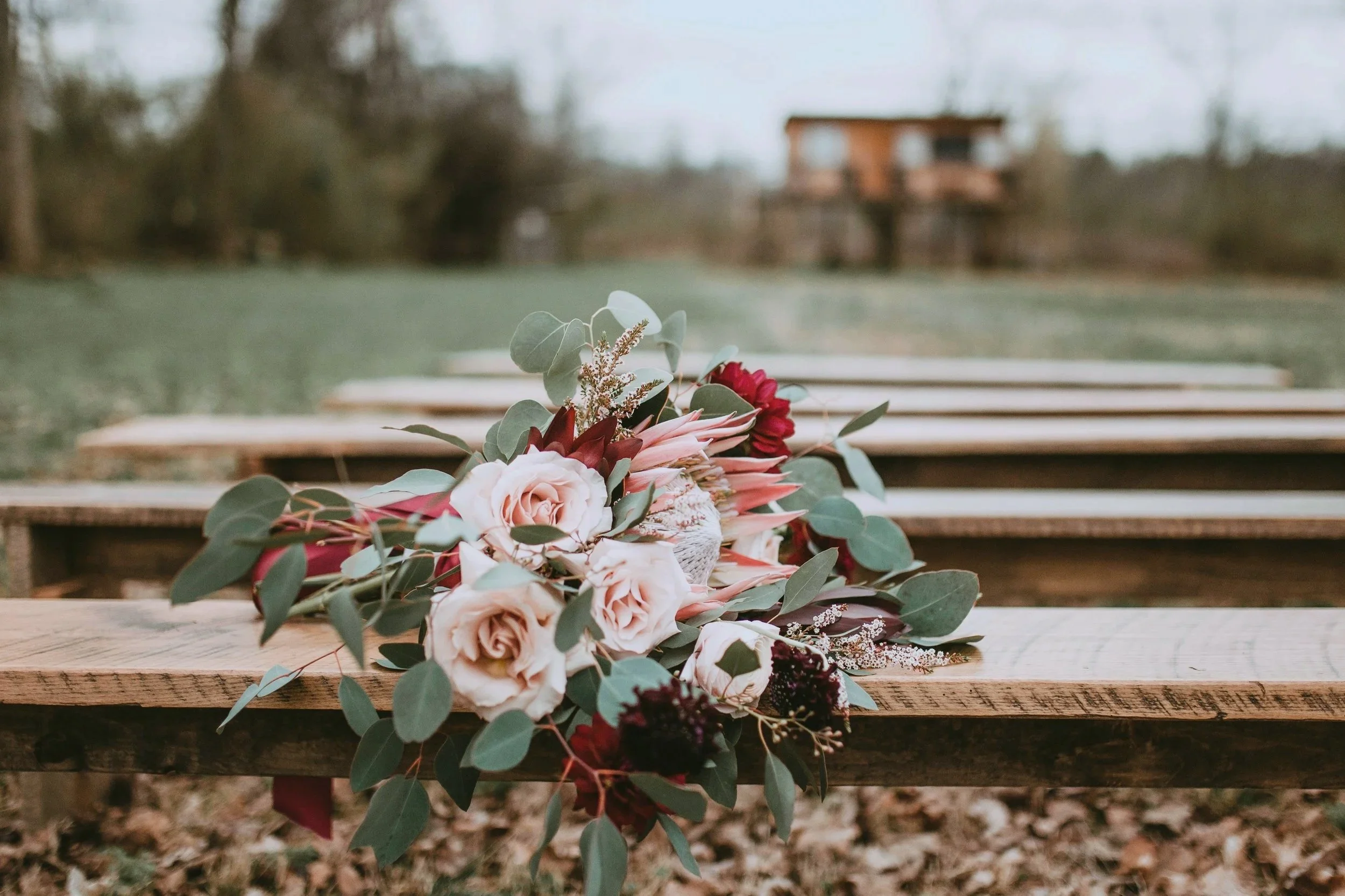 A bouquet of flowers resting on a wooden bench outdoors, with a blurred background of trees and a building.