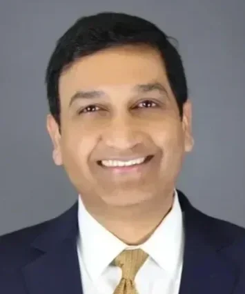 A professional headshot of a smiling man in a suit and tie against a gray background.