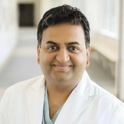 A man in a white doctor's coat smiling in a hospital or clinic hallway.