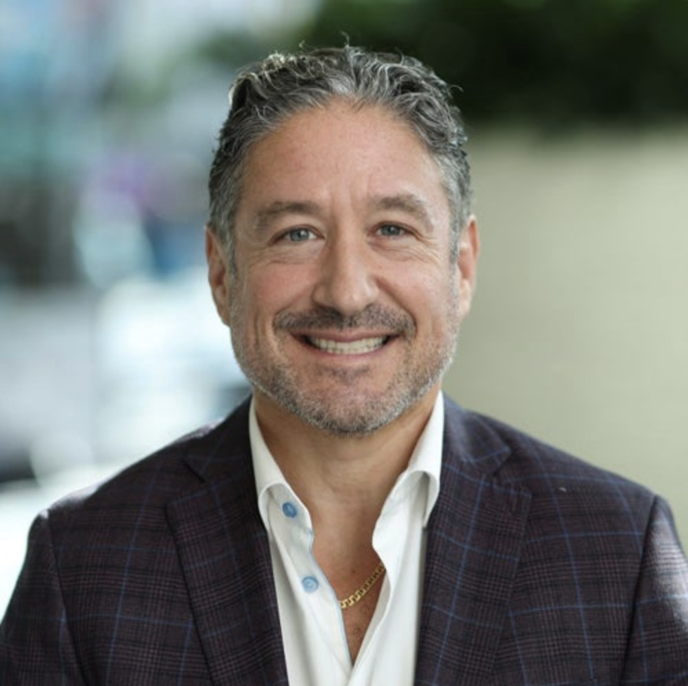 A middle-aged man with gray hair and a beard, wearing a white shirt and a patterned blazer, smiling at the camera.