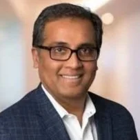 A smiling man wearing glasses, a blue checkered suit, and a white shirt, standing in an indoor setting.