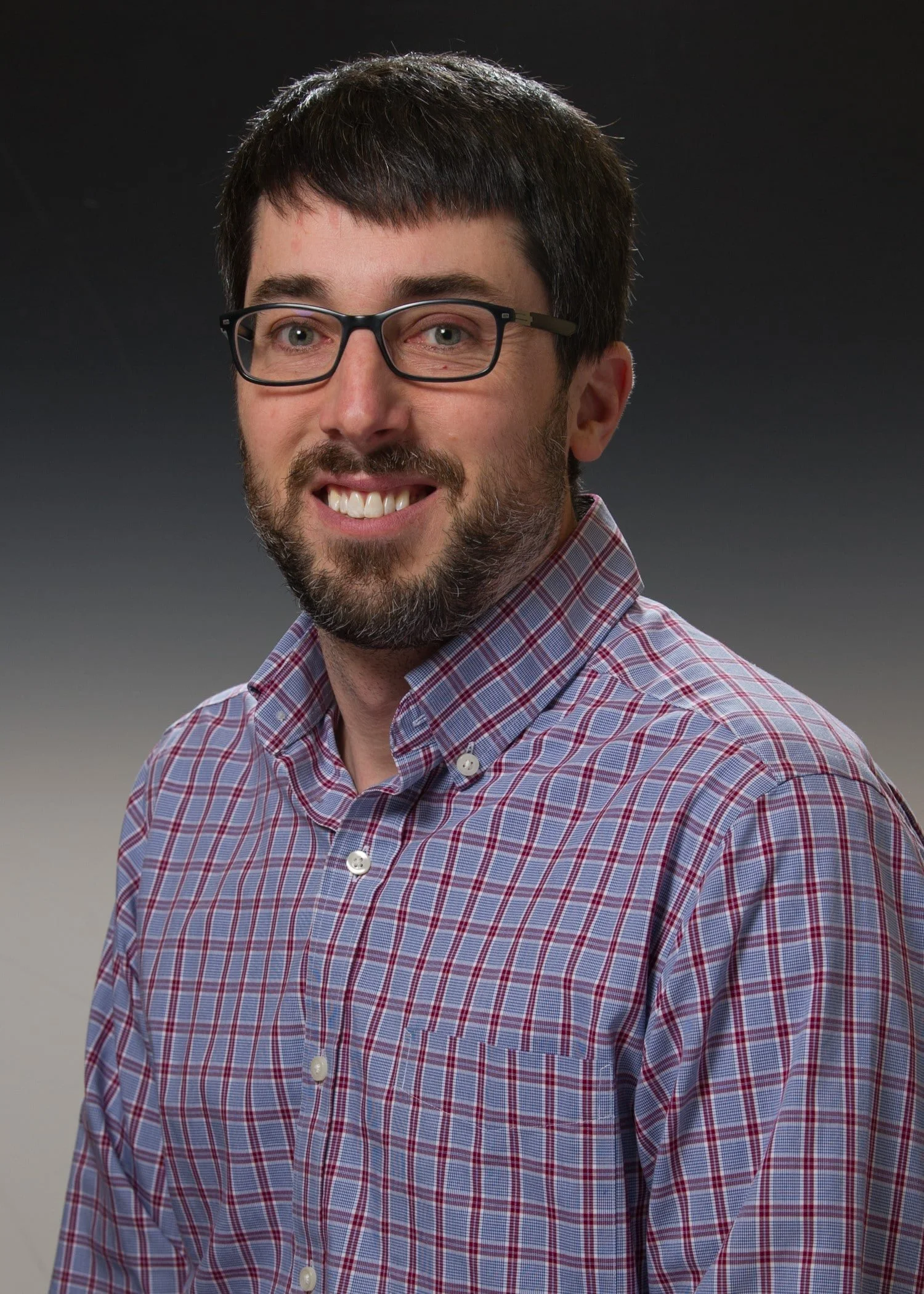 Portrait of a man with dark hair and glasses, smiling, wearing a plaid button-up shirt, set against a gradient background.
