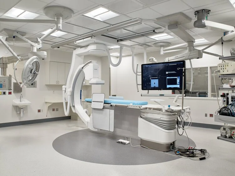 A modern medical operating room with a large C-arm X-ray machine positioned over an operating table, accompanied by a monitor displaying medical imaging, and various medical equipment and machinery around the room.