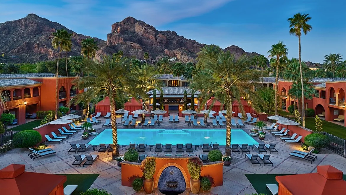 Luxury hotel courtyard with swimming pool, surrounded by palm trees, lounge chairs, umbrellas, and a mountain backdrop at dusk.