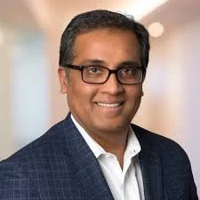 Headshot of a smiling man wearing glasses, a white shirt, and a dark blazer indoors.