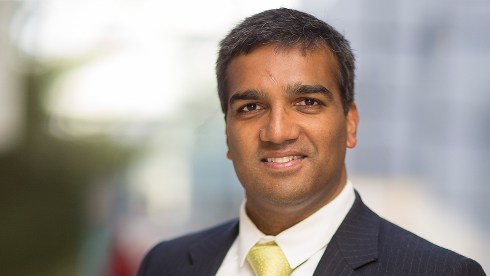 Professional portrait of a man in a dark suit, white shirt, and yellow tie, smiling in an office environment.